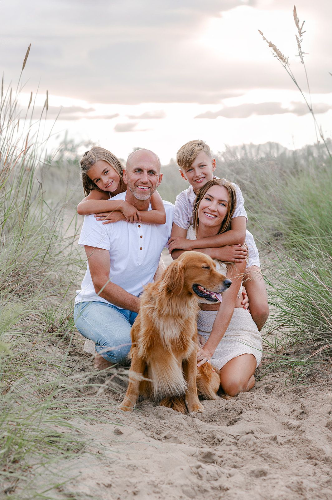 Family of four with golden retriever posing on sandy beach dunes at sunset in white outfits.