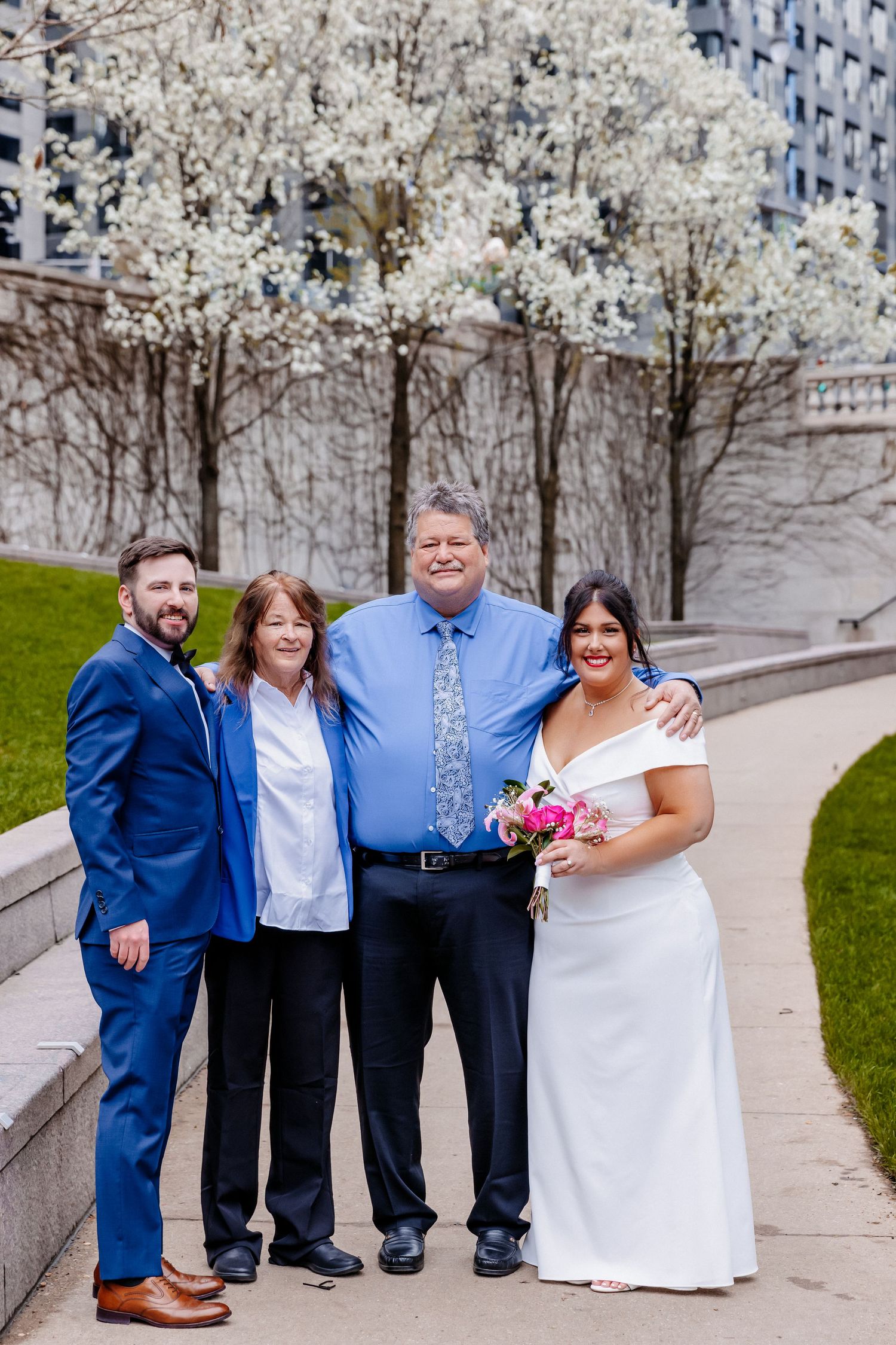 Wedding party of four poses near blooming white trees in urban park, bride holding pink bouquet.