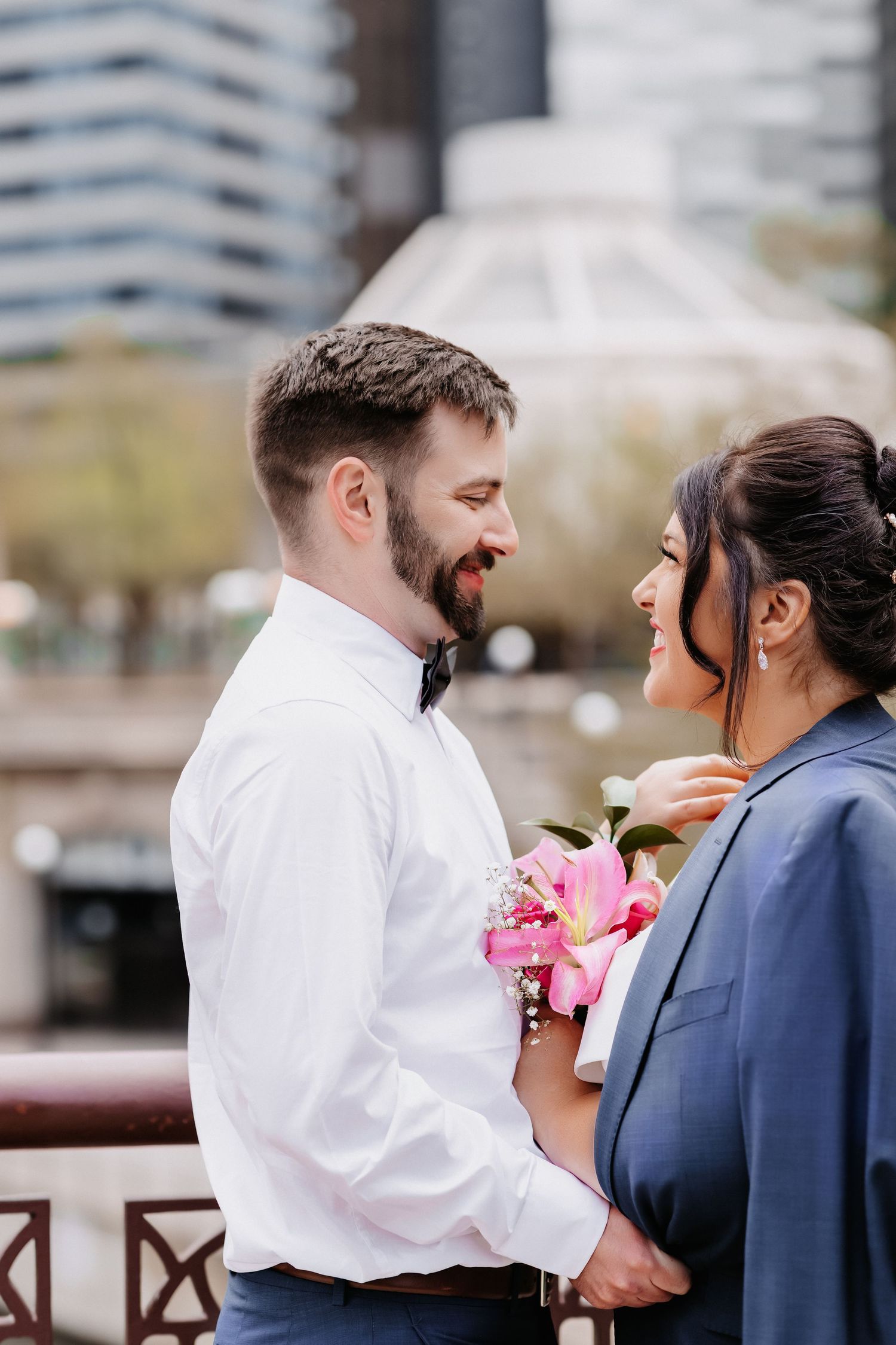 Smiling couple in wedding attire face each other on city bridge, bride holds pink lily bouquet, groom in white shirt.