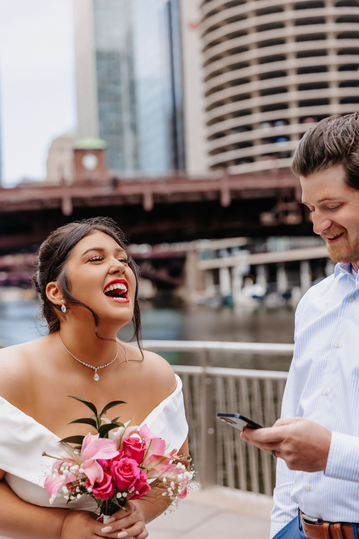 Laughing bride in white gown holds pink roses bouquet beside smiling groom on Chicago Riverwalk.