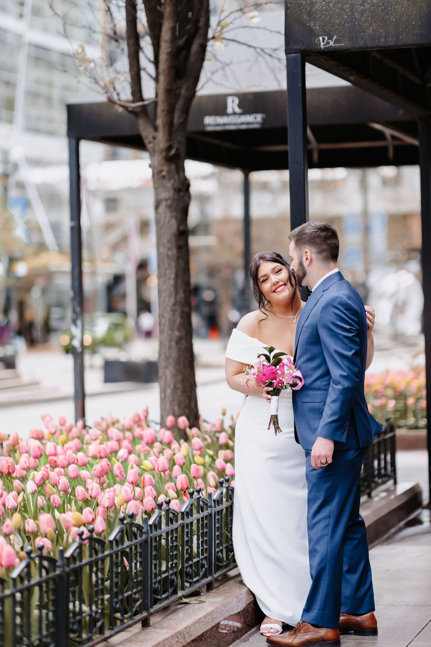 Wedding couple embraces near Renaissance hotel entrance surrounded by blooming pink tulips in spring.