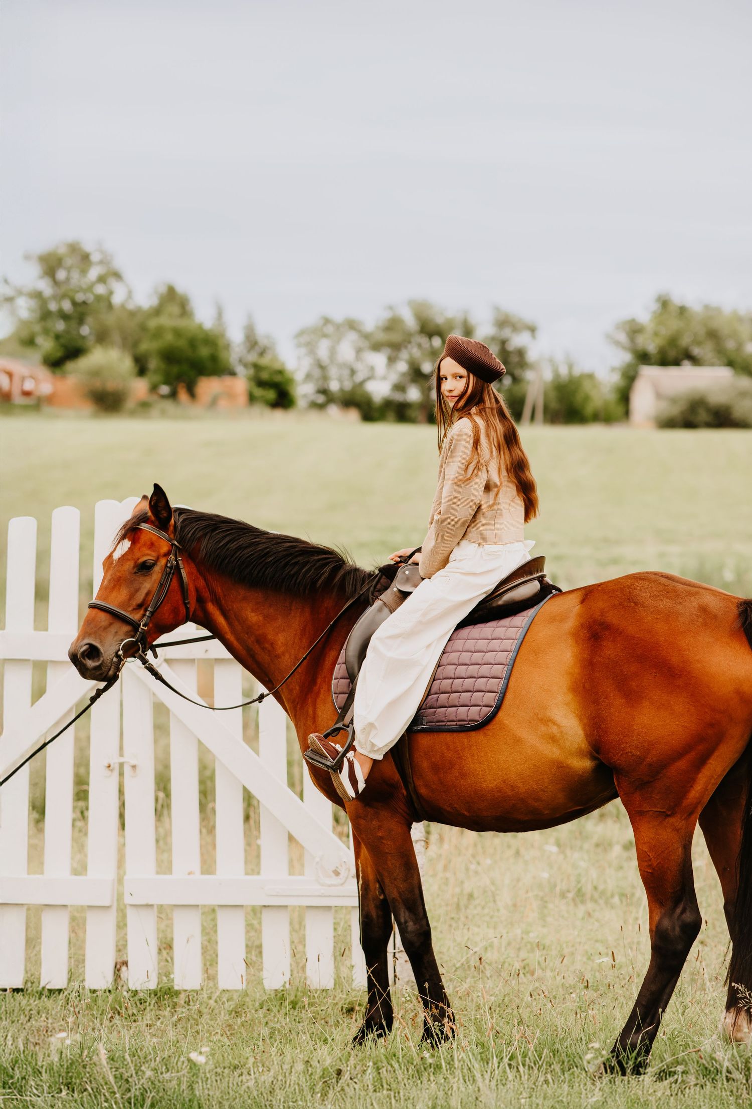 Young woman with long hair riding a chestnut horse by a white picket fence.