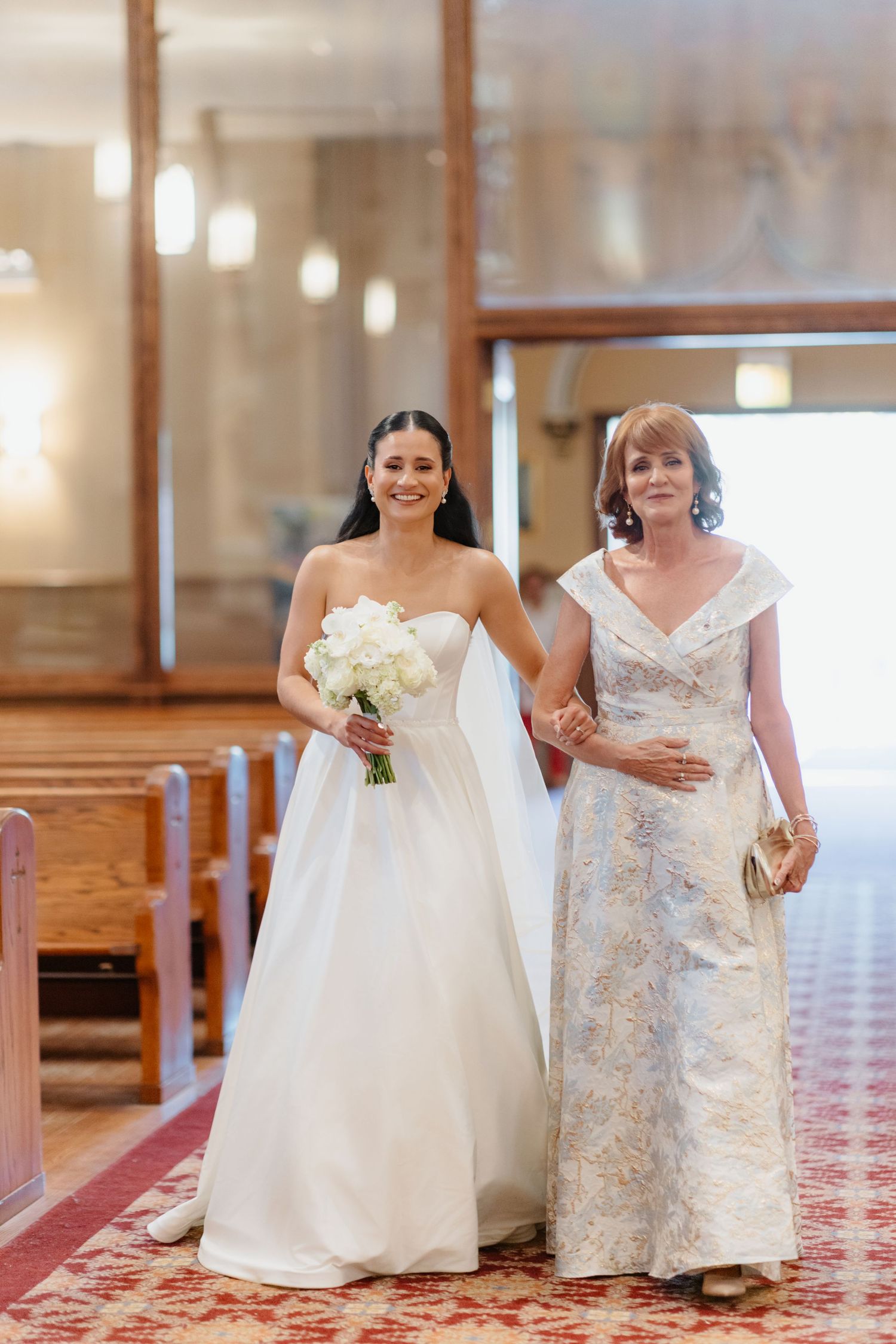 Bride in strapless white gown walks down church aisle with mother during wedding ceremony.