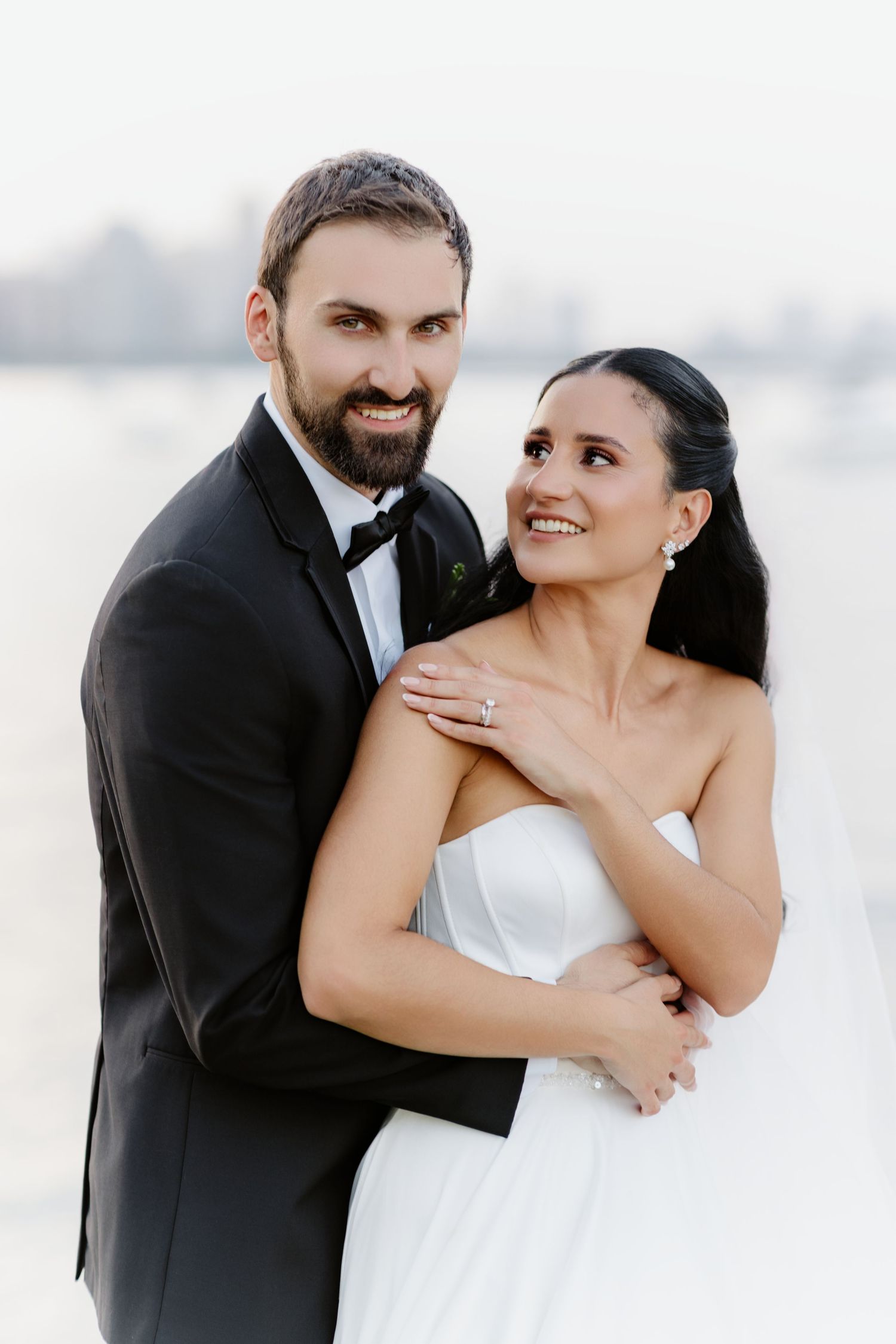 Groom embraces bride from behind as both smile warmly in a soft, dreamy outdoor wedding portrait.