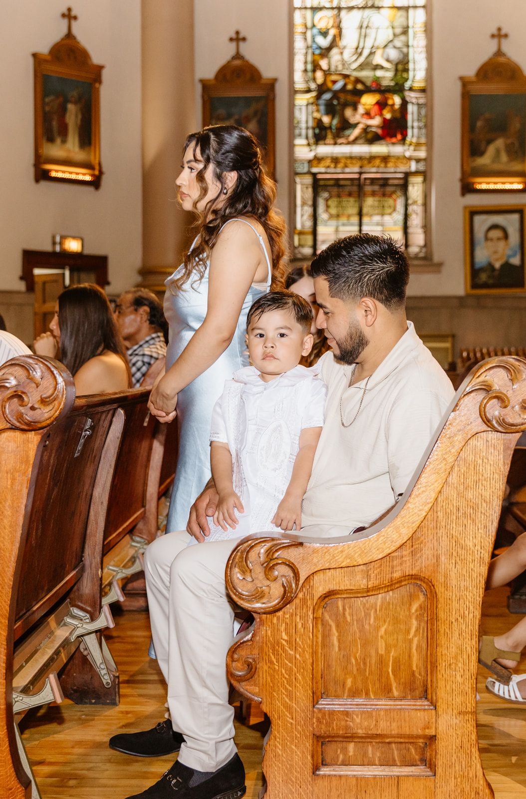 Family sitting in wooden church pews during a baptism ceremony, with stained glass windows in the background.