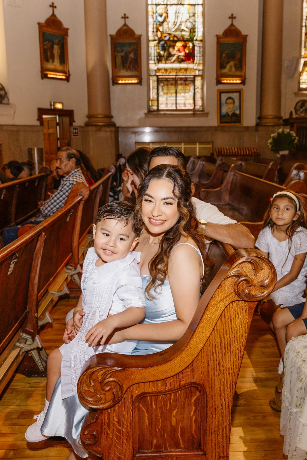 A woman and toddler in white sit smiling on a wooden church pew during a religious ceremony.