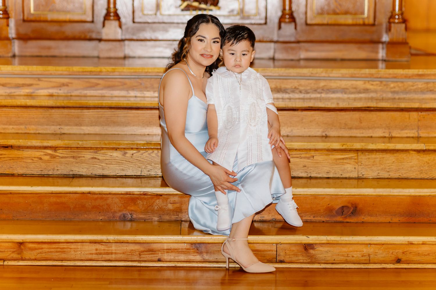 Mother in blue dress sitting on wooden church steps holding baby in white baptism outfit.