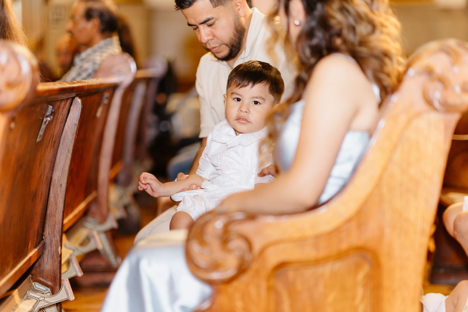 A young child in white sits on a church pew with family during a wedding ceremony.