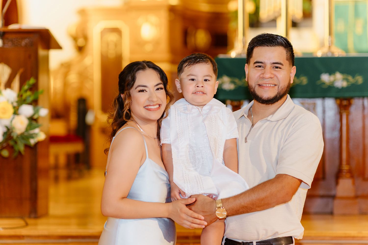 A smiling couple holds their baby dressed in white inside a warmly lit church during a baptism ceremony.