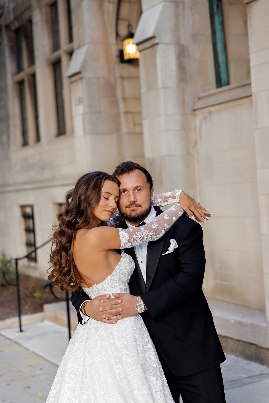 Bride in open-back lace wedding dress and groom in black tuxedo share romantic embrace outdoors.