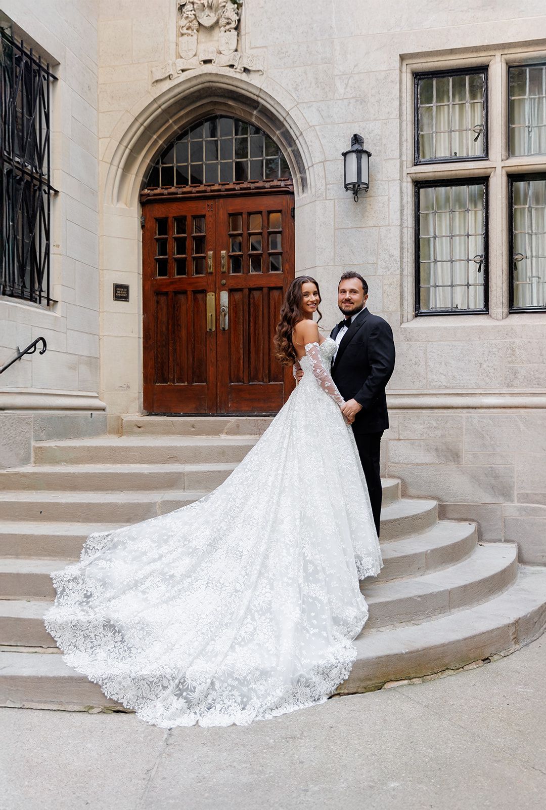 Bride in lace ball gown with long train and groom in tuxedo posing on stone steps before ornate wooden doors.