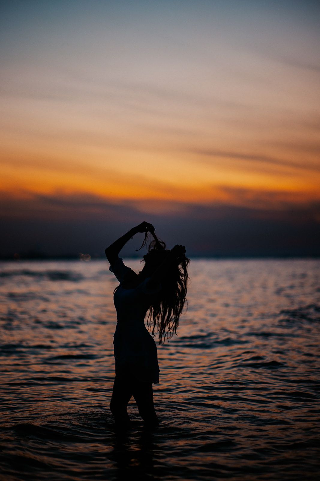 Silhouette of a woman standing in ocean water at sunset, with vibrant orange and purple sky reflecting on the sea.