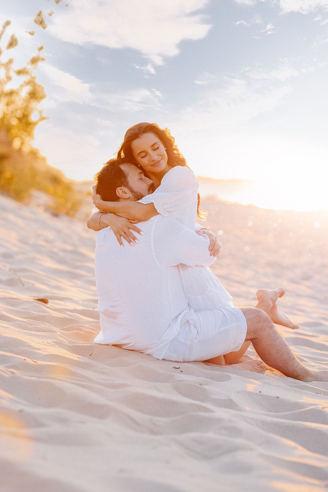 Couple embracing on sandy beach dunes bathed in warm golden sunset light.