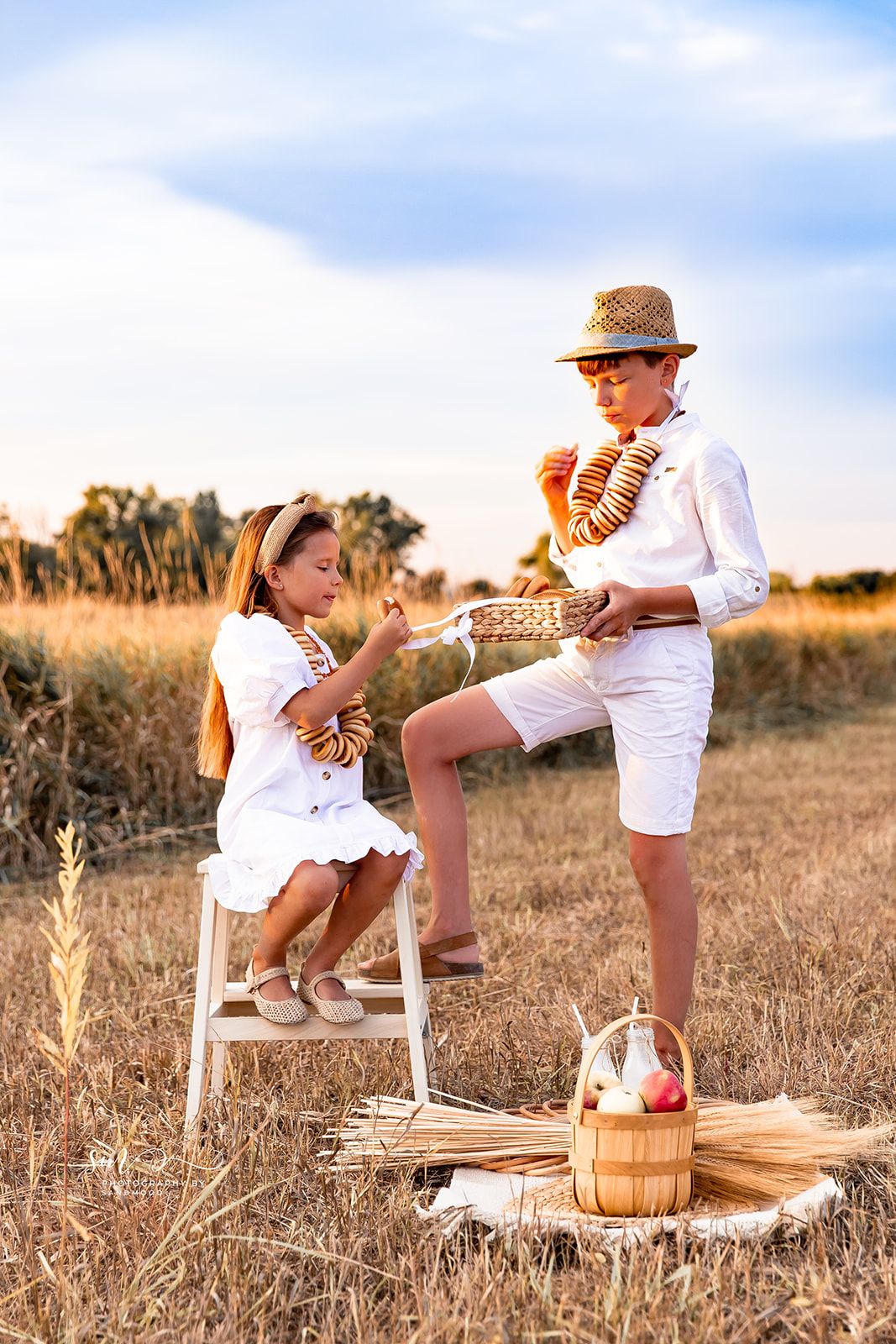 Two children in white outfits enjoy a picnic in a golden field at sunset, sharing bread from a wicker basket.