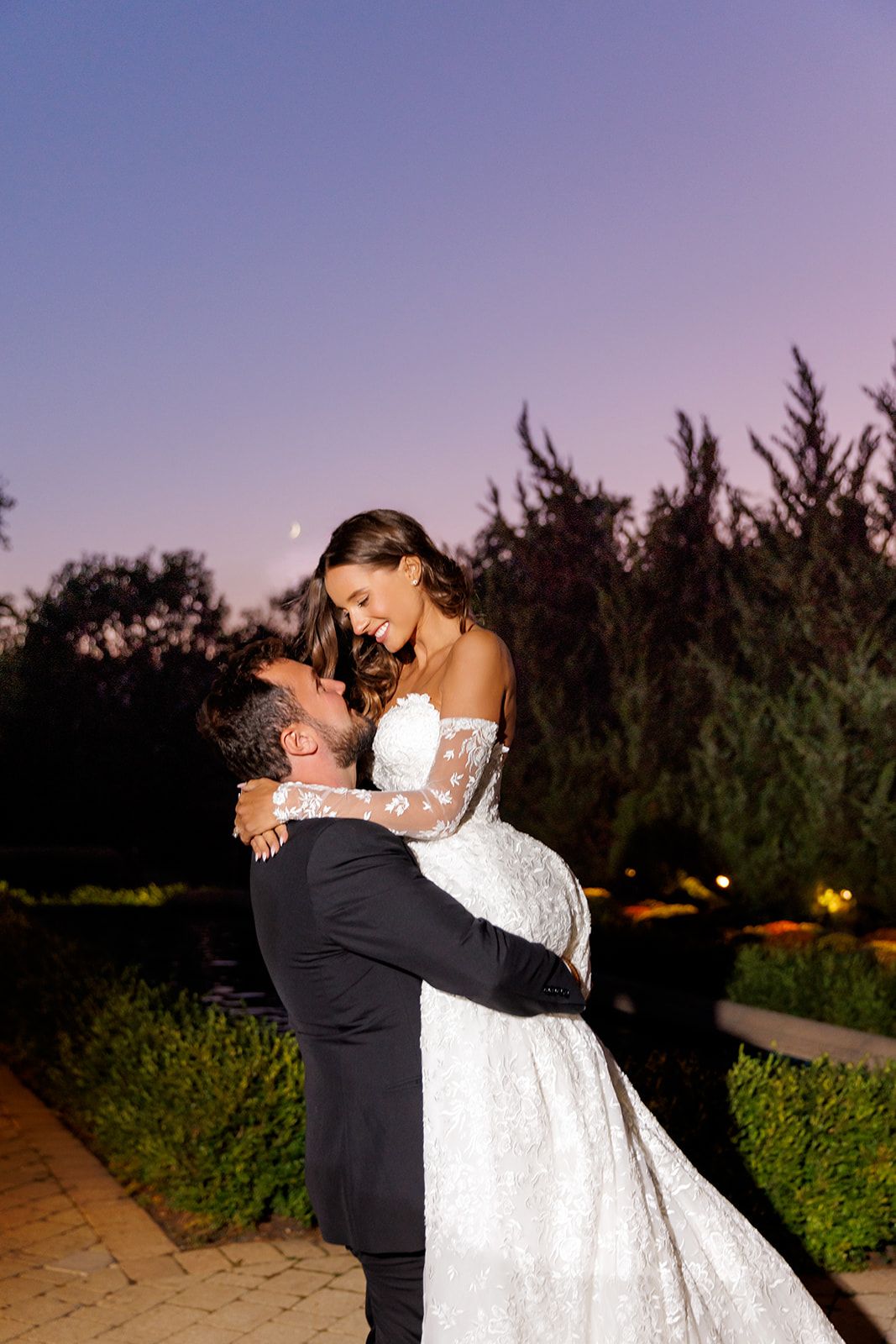 Bride in lace gown lifted by groom in tuxedo, smiling joyfully under a purple dusk sky.