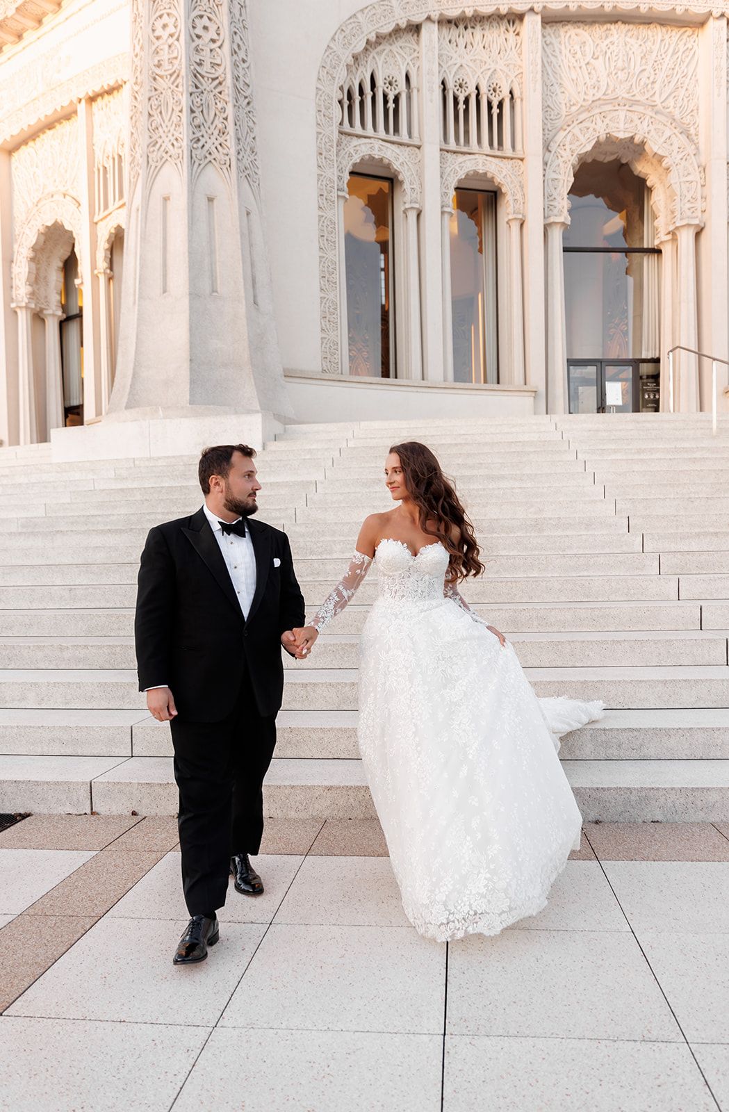 Bride in lace gown and groom in tuxedo walk hand-in-hand before an ornate white building.