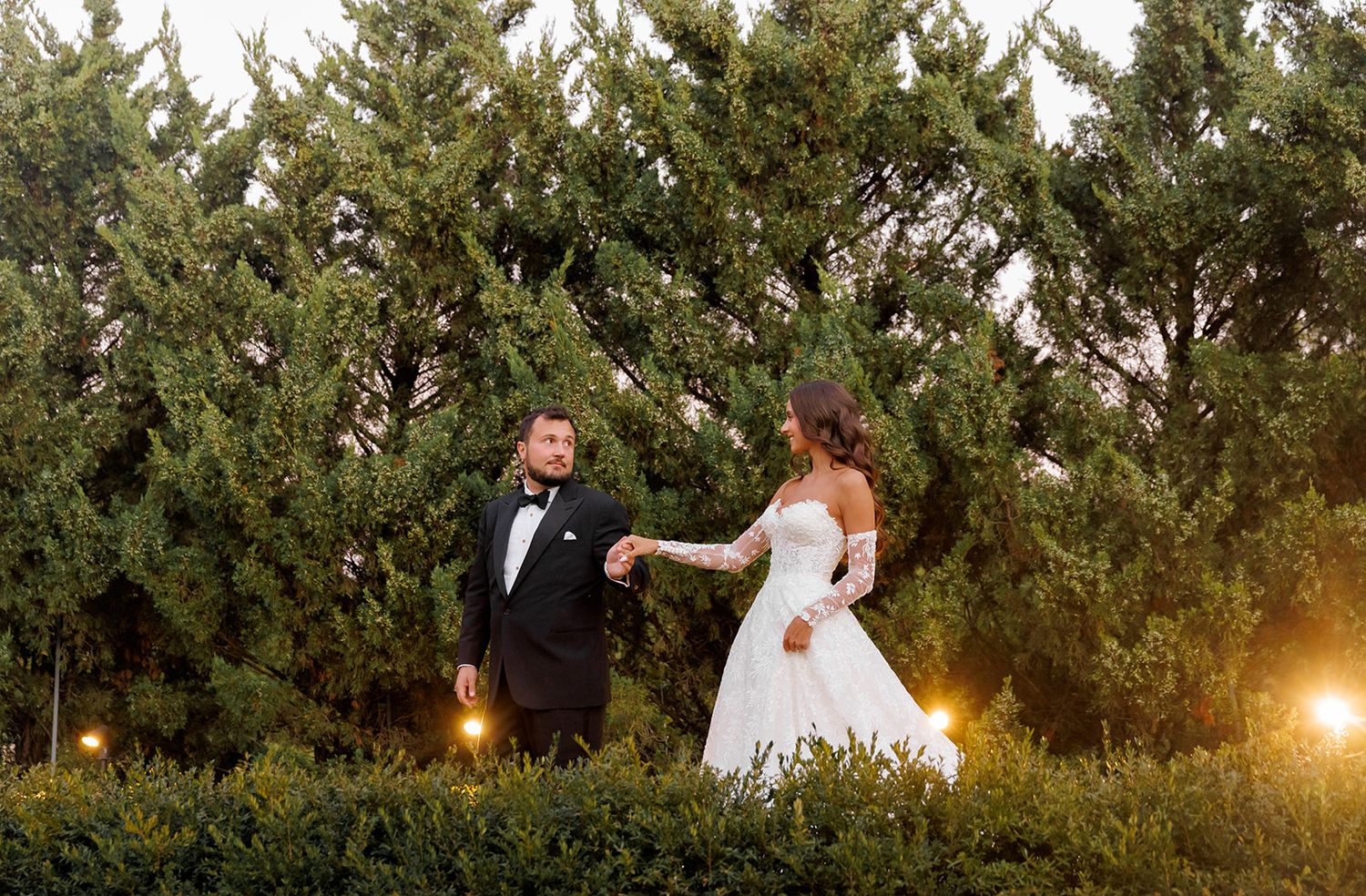 Bride and groom hold hands in front of lush green trees at dusk, bride in white gown, groom in black tuxedo.
