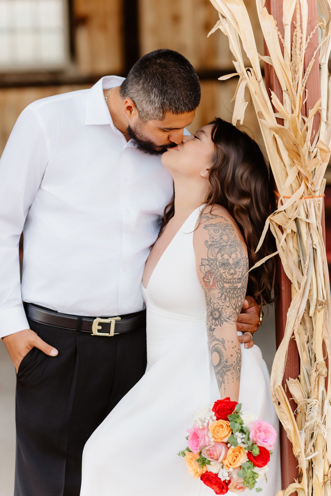 Bride and groom share a kiss near dried corn stalks, bride holding colorful bouquet wearing white gown with tattoos.
