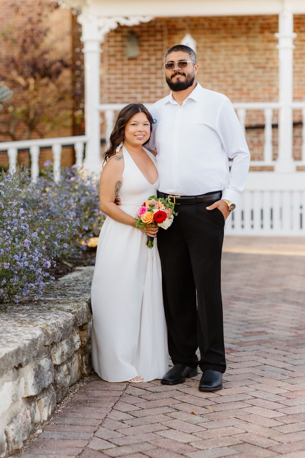 Smiling bride in white dress and groom in white shirt holding colorful bouquet outside brick building.