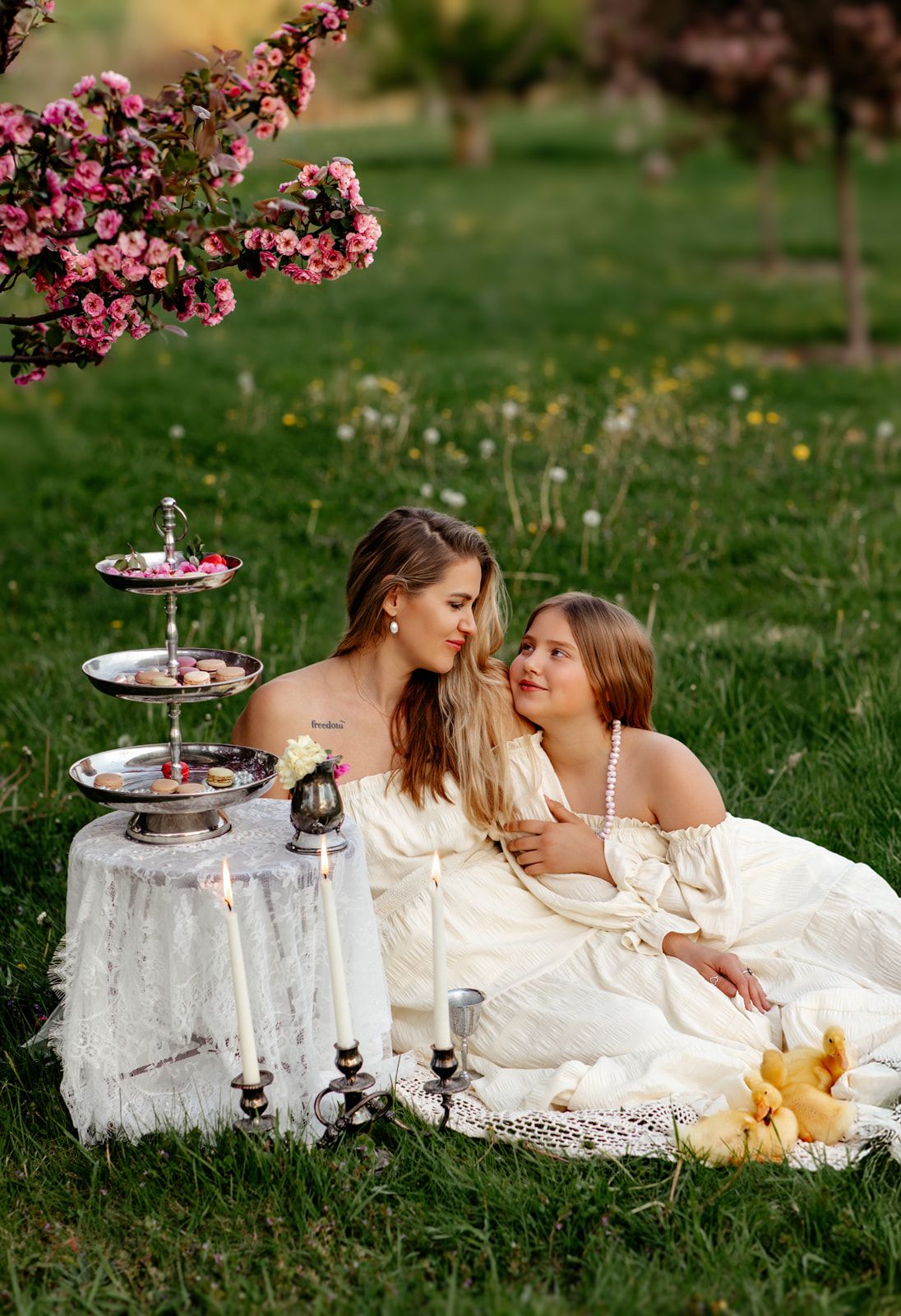 Mother and daughter in cream dresses enjoy romantic garden picnic with candles and tiered stand under pink blooms.
