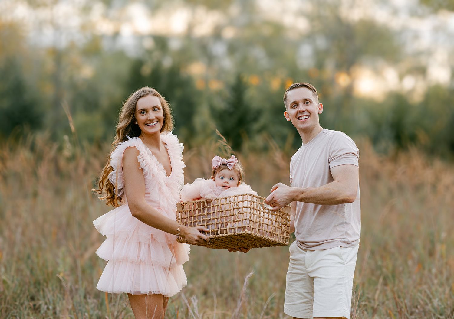 A young family smiles outdoors, holding a wicker basket with their baby girl in a pink bow in a sunny field.