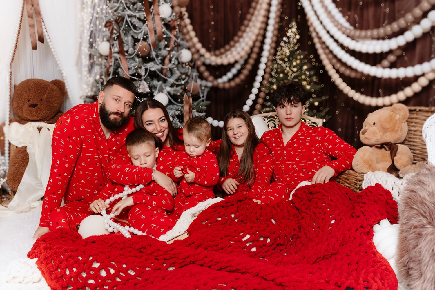 Large family wearing matching red pajamas sits together on a bed surrounded by Christmas trees and teddy bears.