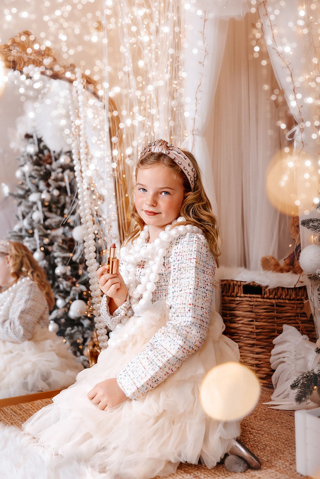 Young girl in white dress and sparkly headband poses near Christmas tree with festive lights and decorations.