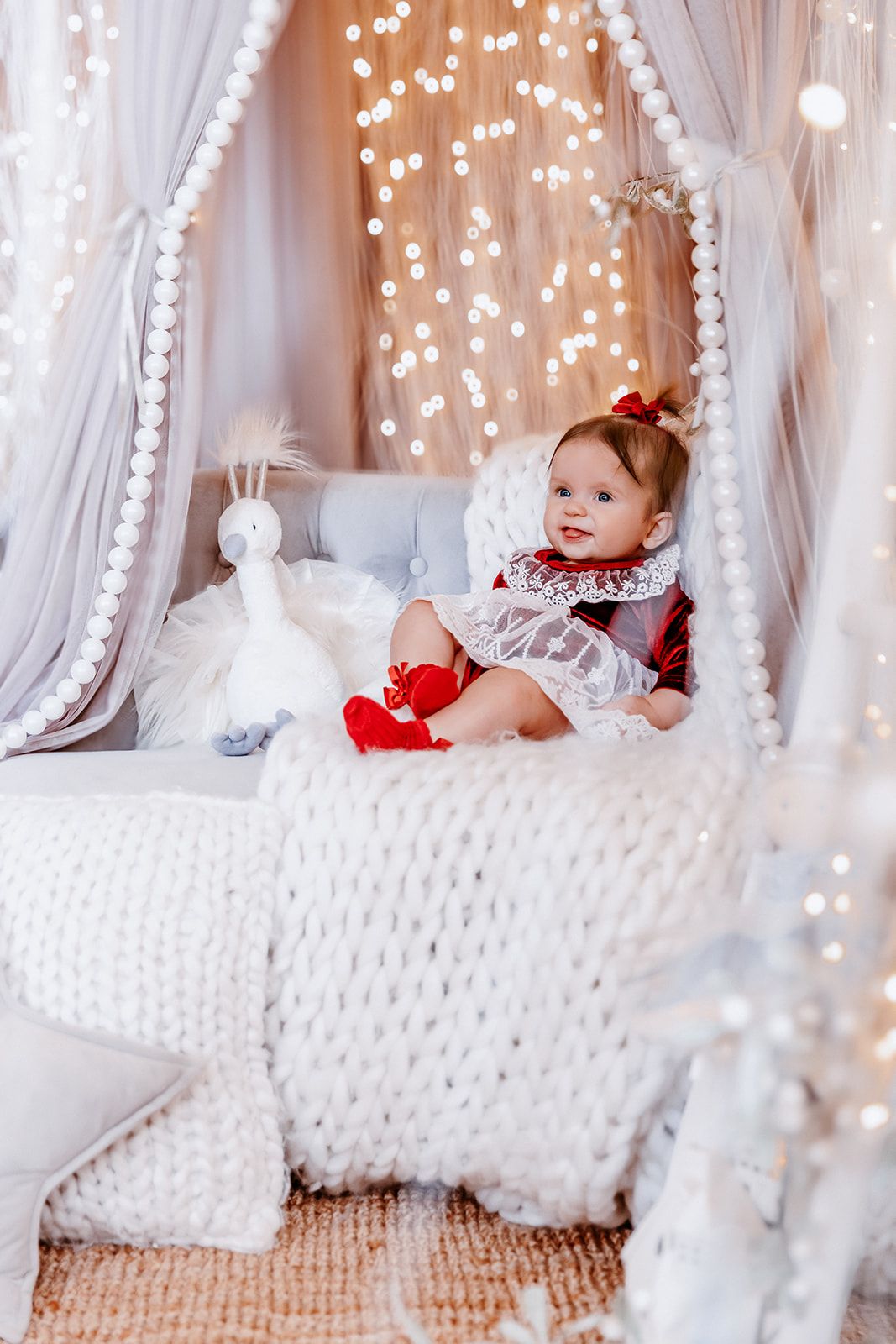 Baby girl in red and white dress sits on a cozy knit blanket in a canopy bed with fairy lights.