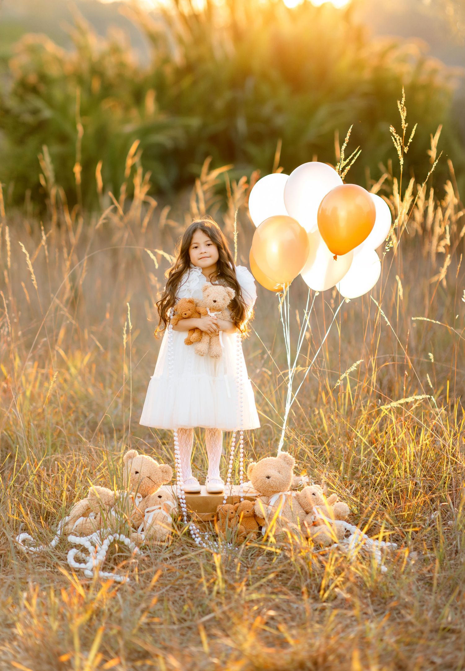 Girl in white dress holds teddy bear surrounded by stuffed animals and gold balloons in a golden field at sunset.