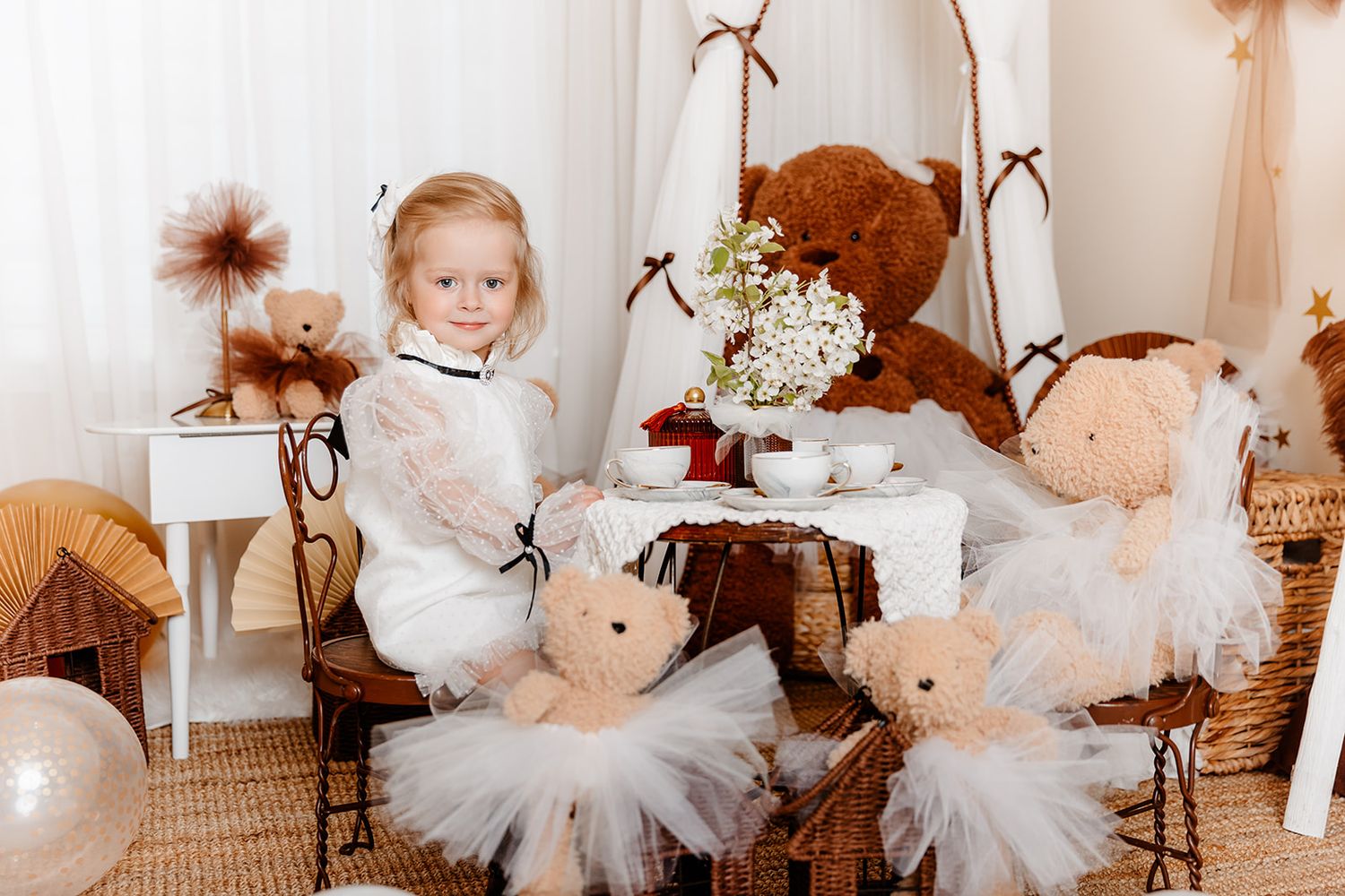 Young girl in white dress poses with teddy bears at a tea party setup with floral decor and wicker furniture.