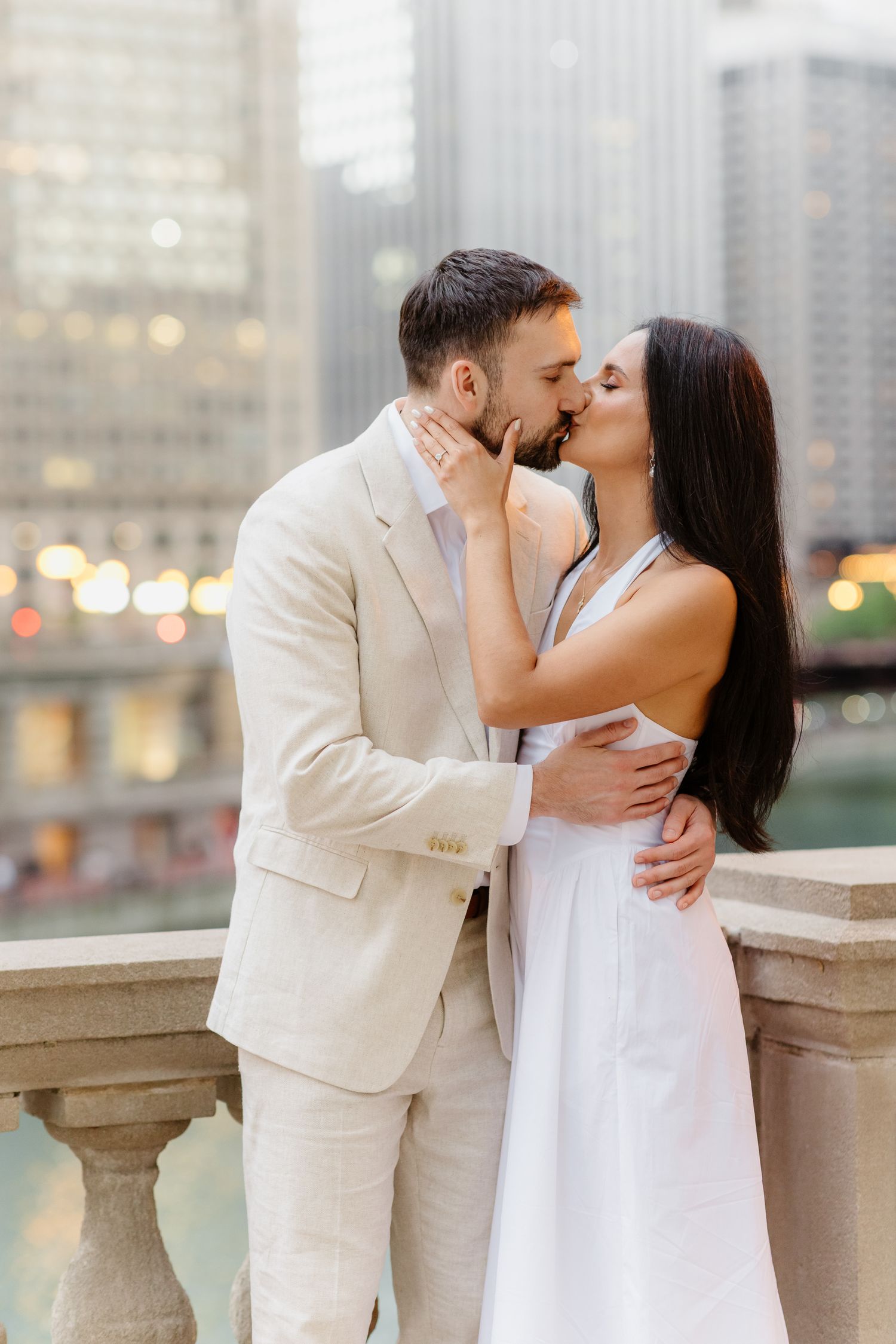 Couple sharing a romantic kiss on a Chicago riverwalk balcony, man in cream suit, woman in white halter dress.