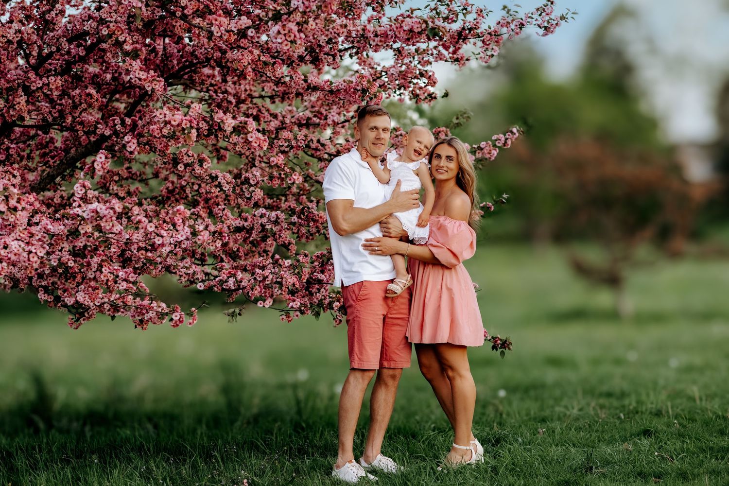 Happy family with baby posing near pink blossoming tree on green lawn in spring park.