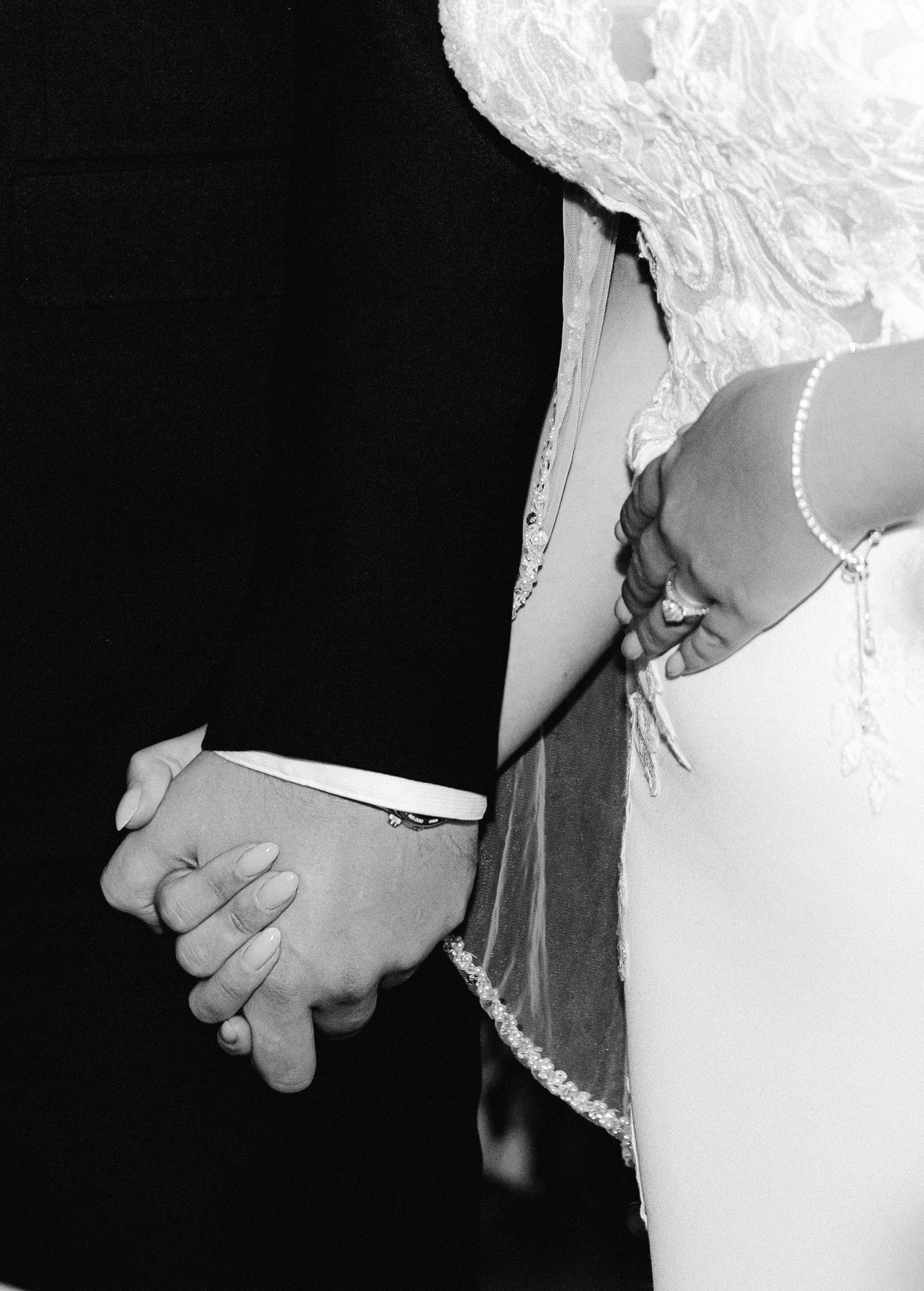 Black and white close-up of bride and groom holding hands showing wedding ring and pearl bracelet details.