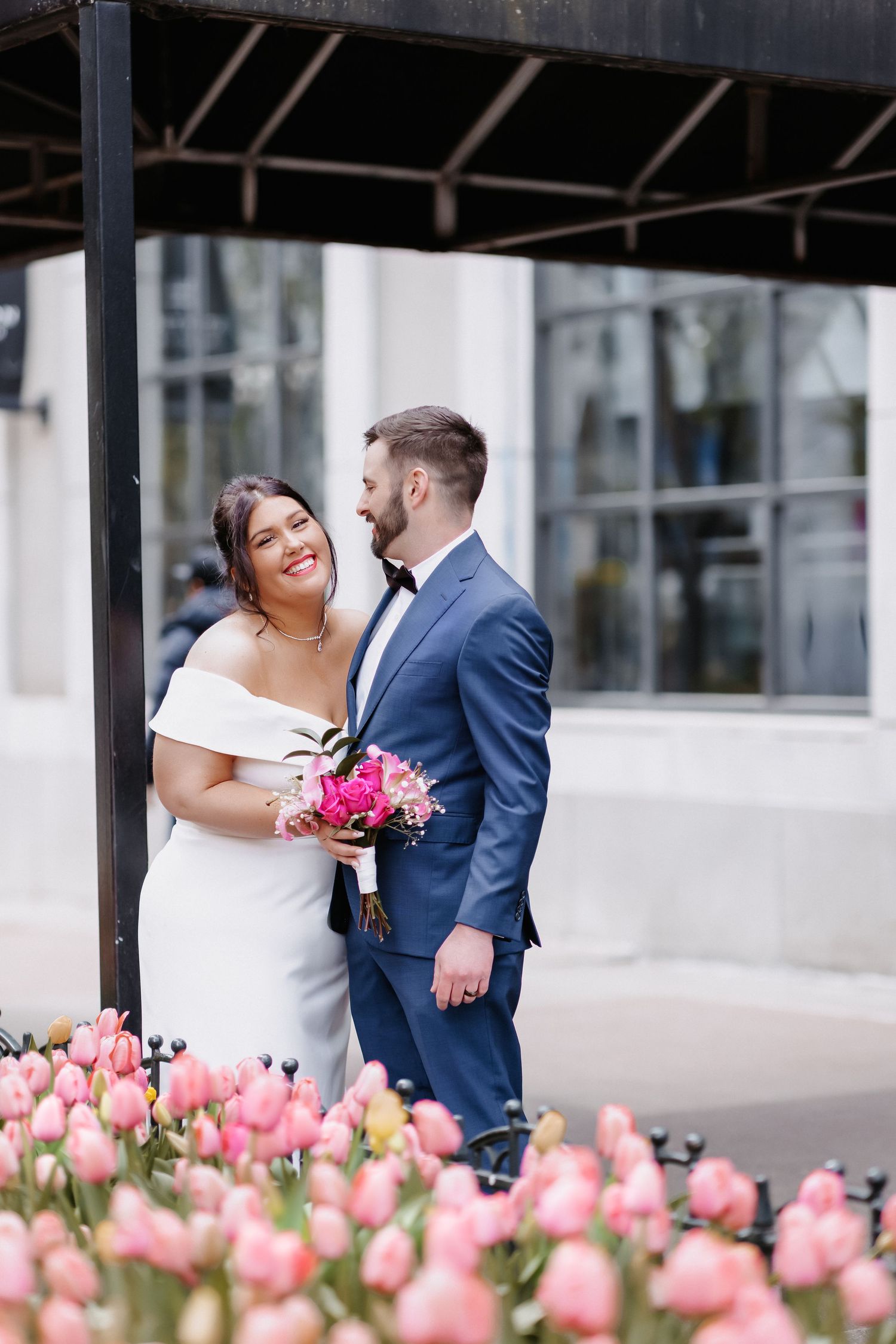 Smiling bride in white off-shoulder gown holds pink bouquet with groom in blue suit near pink tulips.