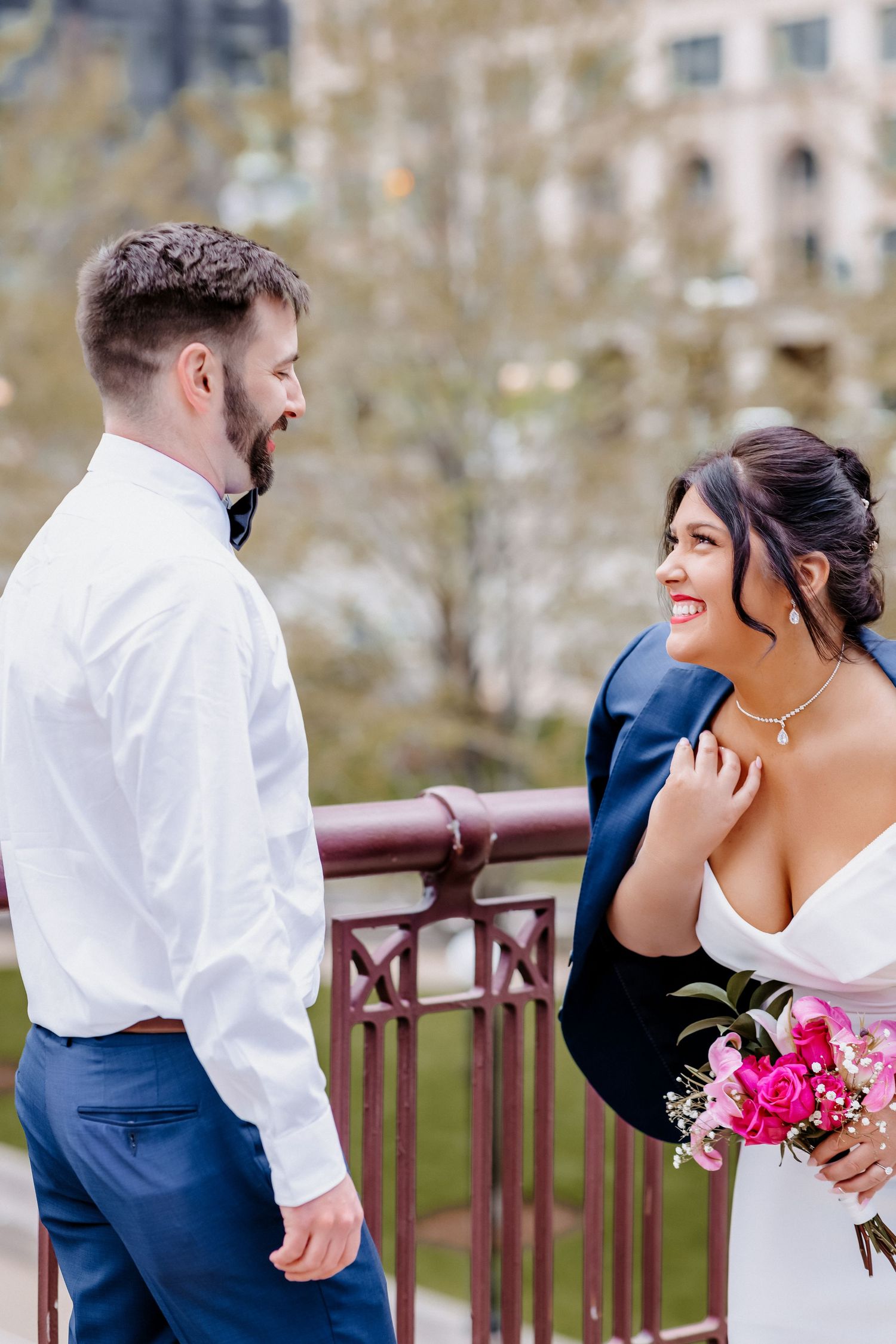 Bride wearing grooms blue jacket laughs with bearded groom in white shirt on ornate bridge in urban setting.