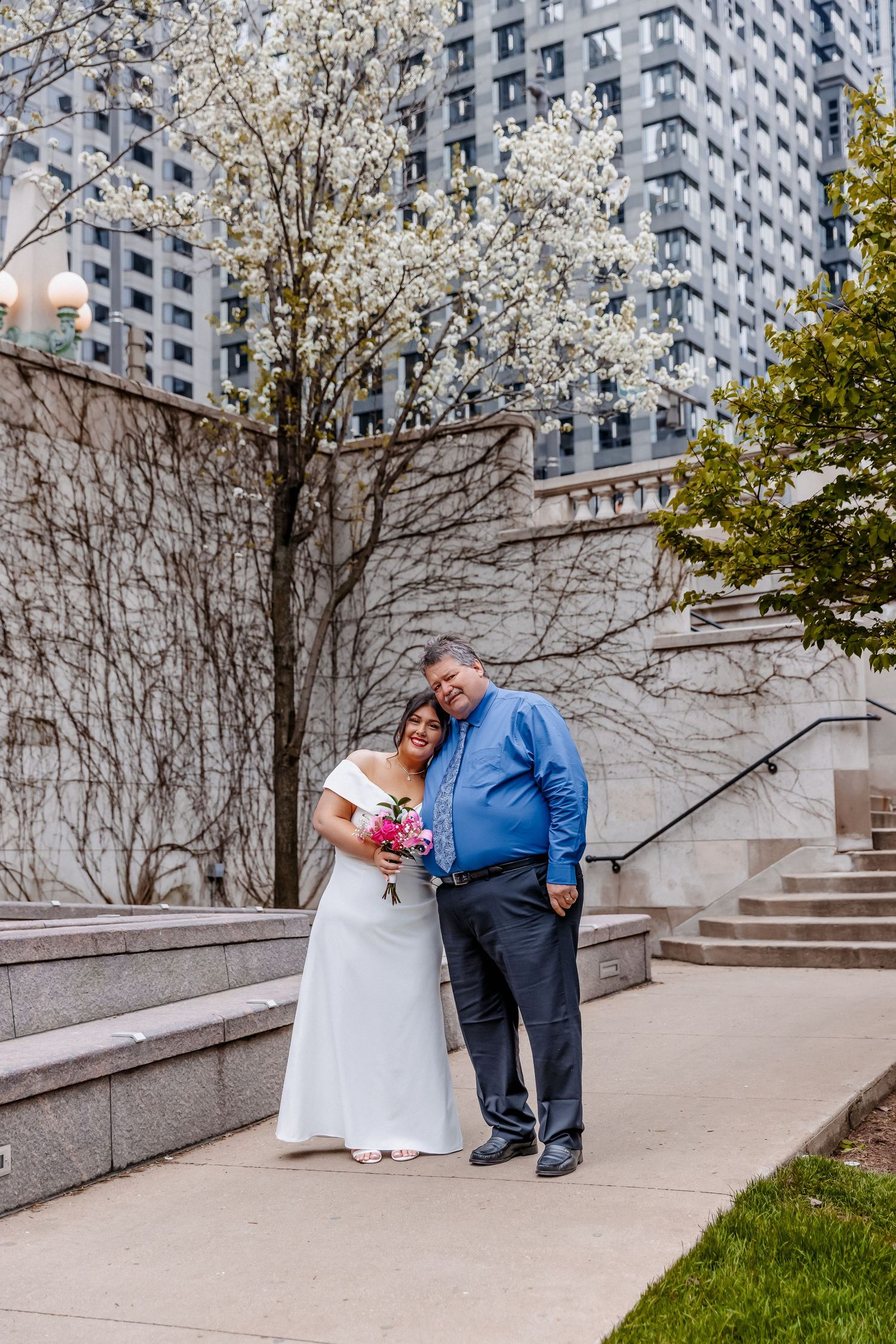 Bride and groom embrace near white flowering trees and stone steps in an urban garden setting.