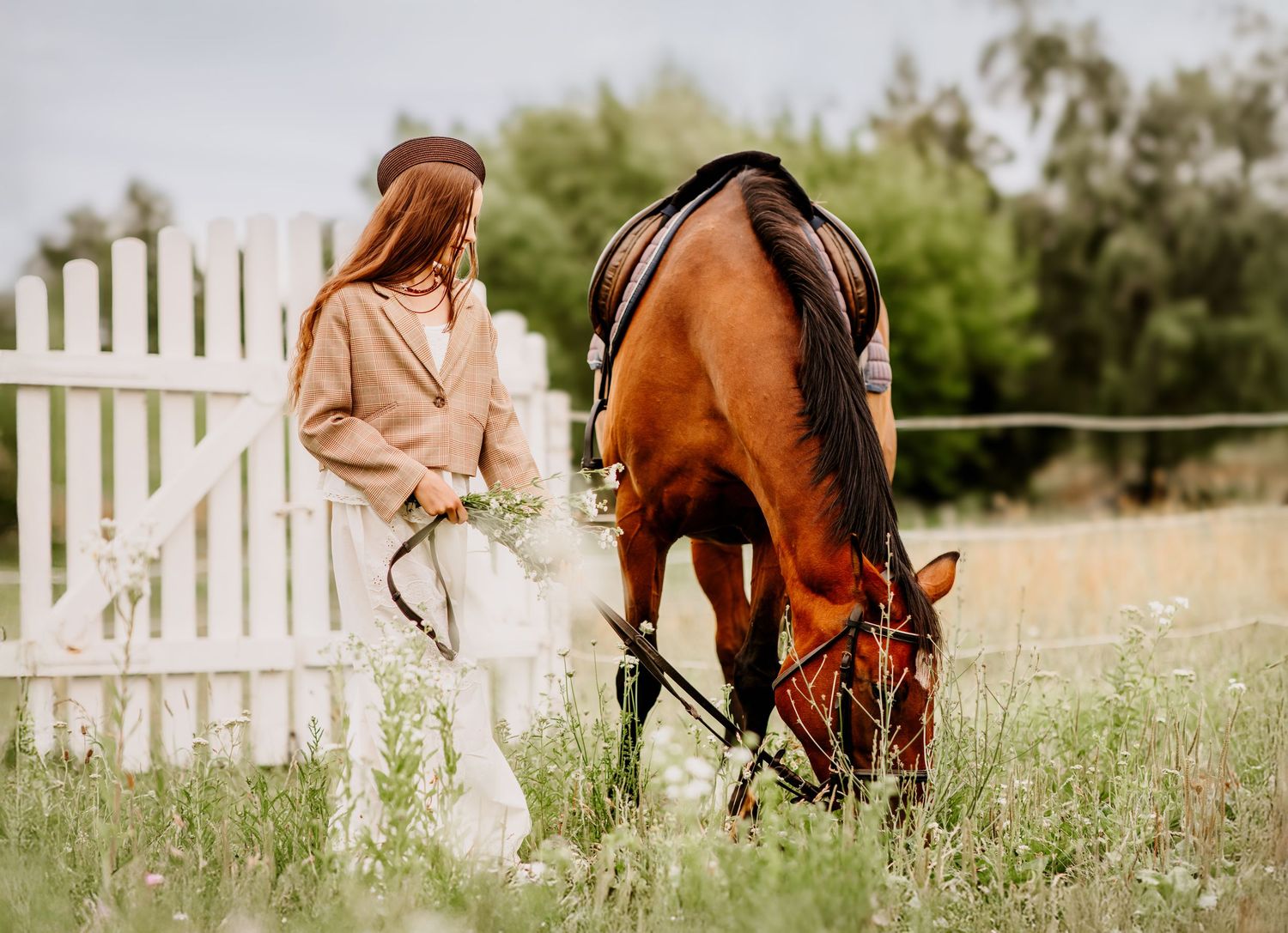 Woman in brown hat leads a bay horse grazing in green meadow beside a white picket fence.