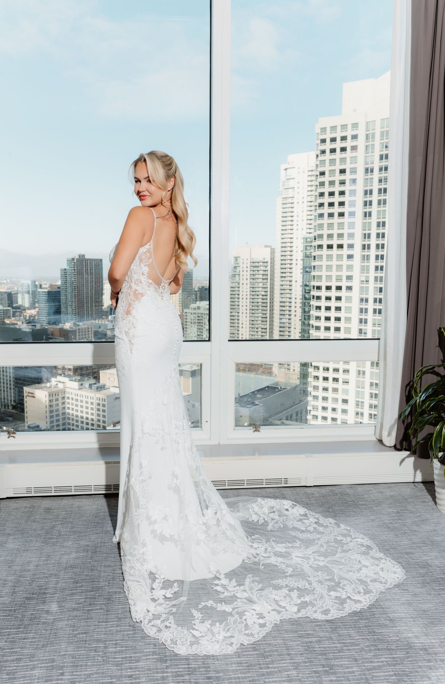 Bride in backless lace wedding gown with long train poses before floor-to-ceiling city view window.