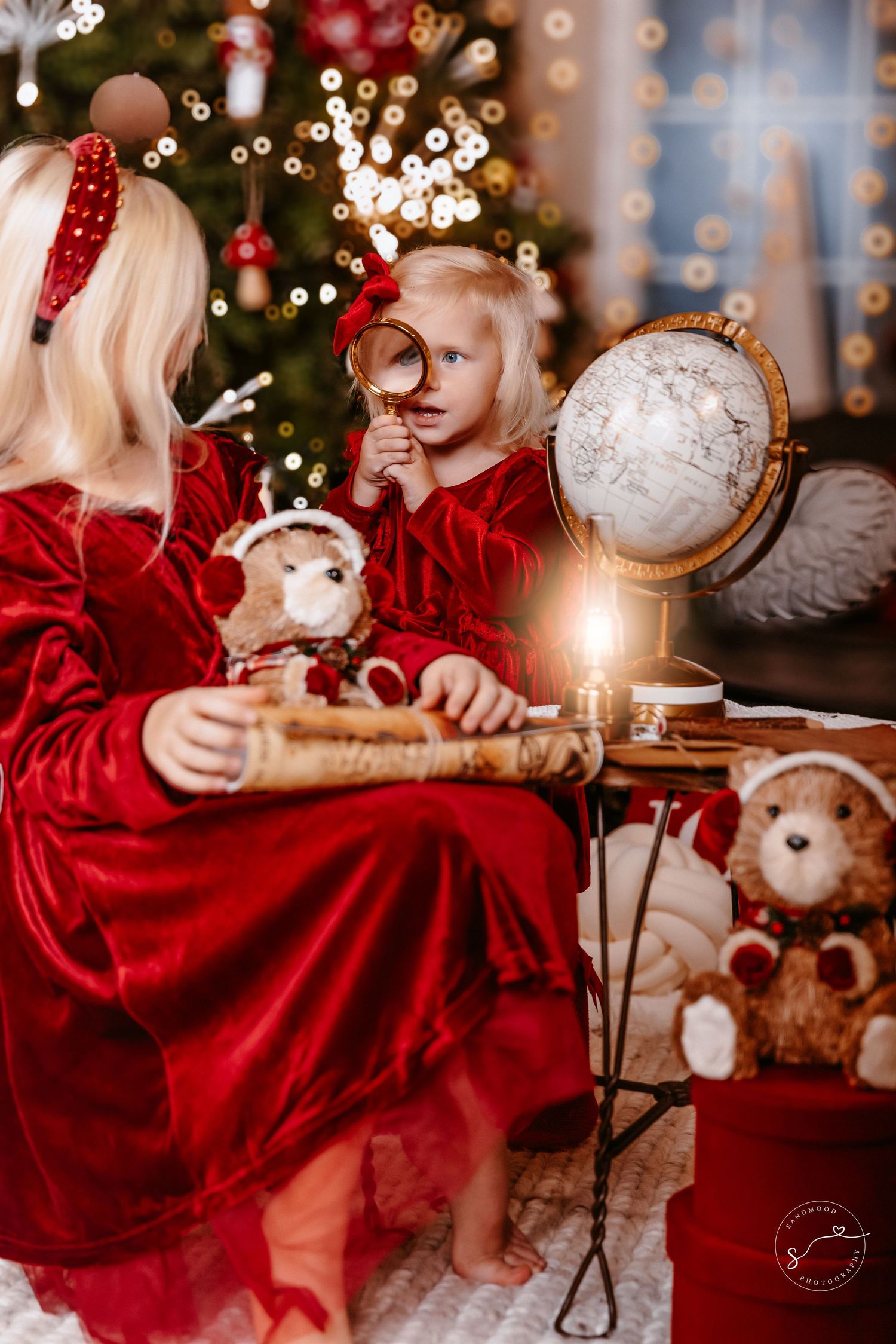 Two blonde girls in red velvet dresses explore globe with magnifying glass by candlelight.