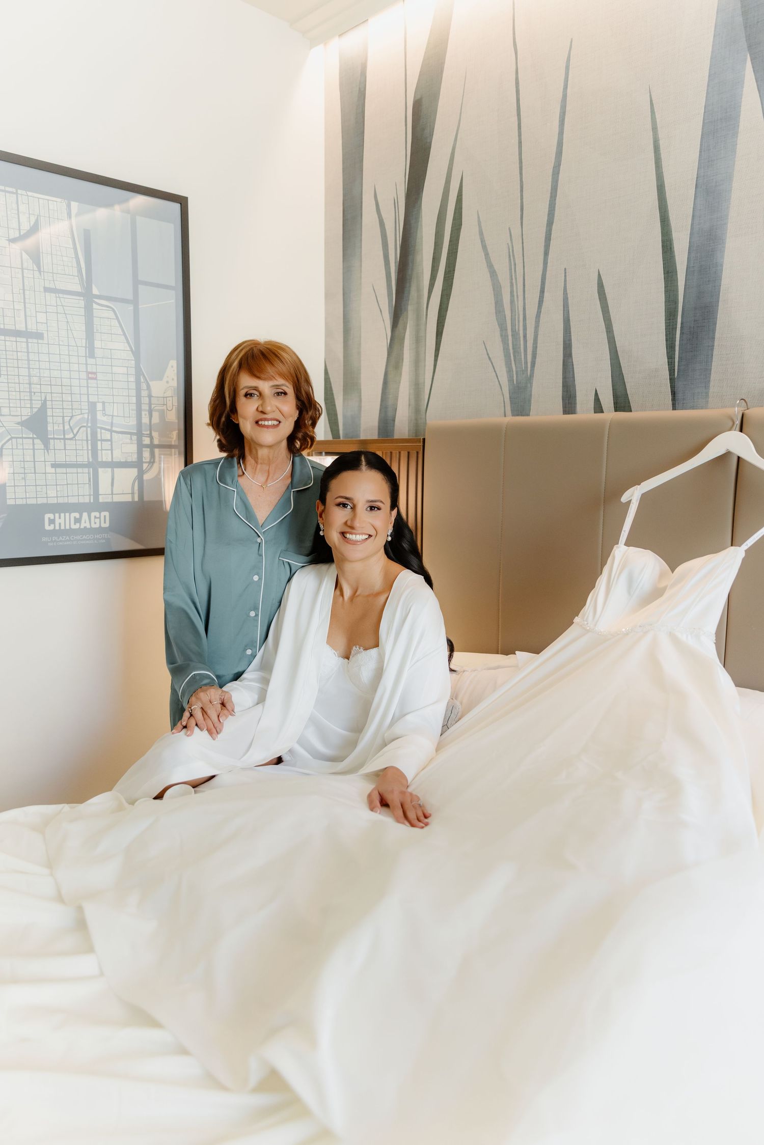 Bride in white robe sits on hotel bed with mother beside hanging wedding gown nearby.