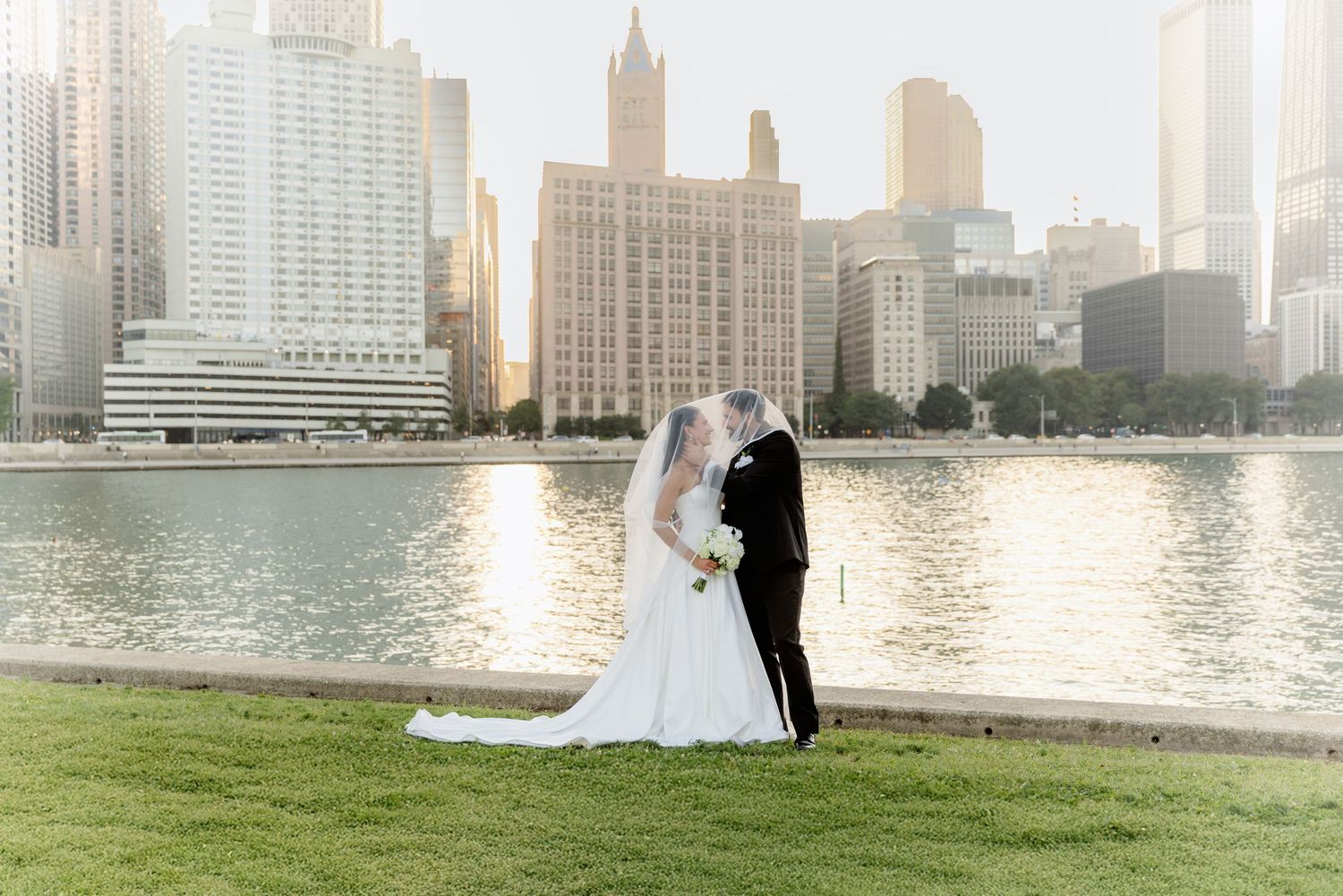Bride and groom share a romantic kiss by the waterfront with a stunning city skyline backdrop at golden hour.