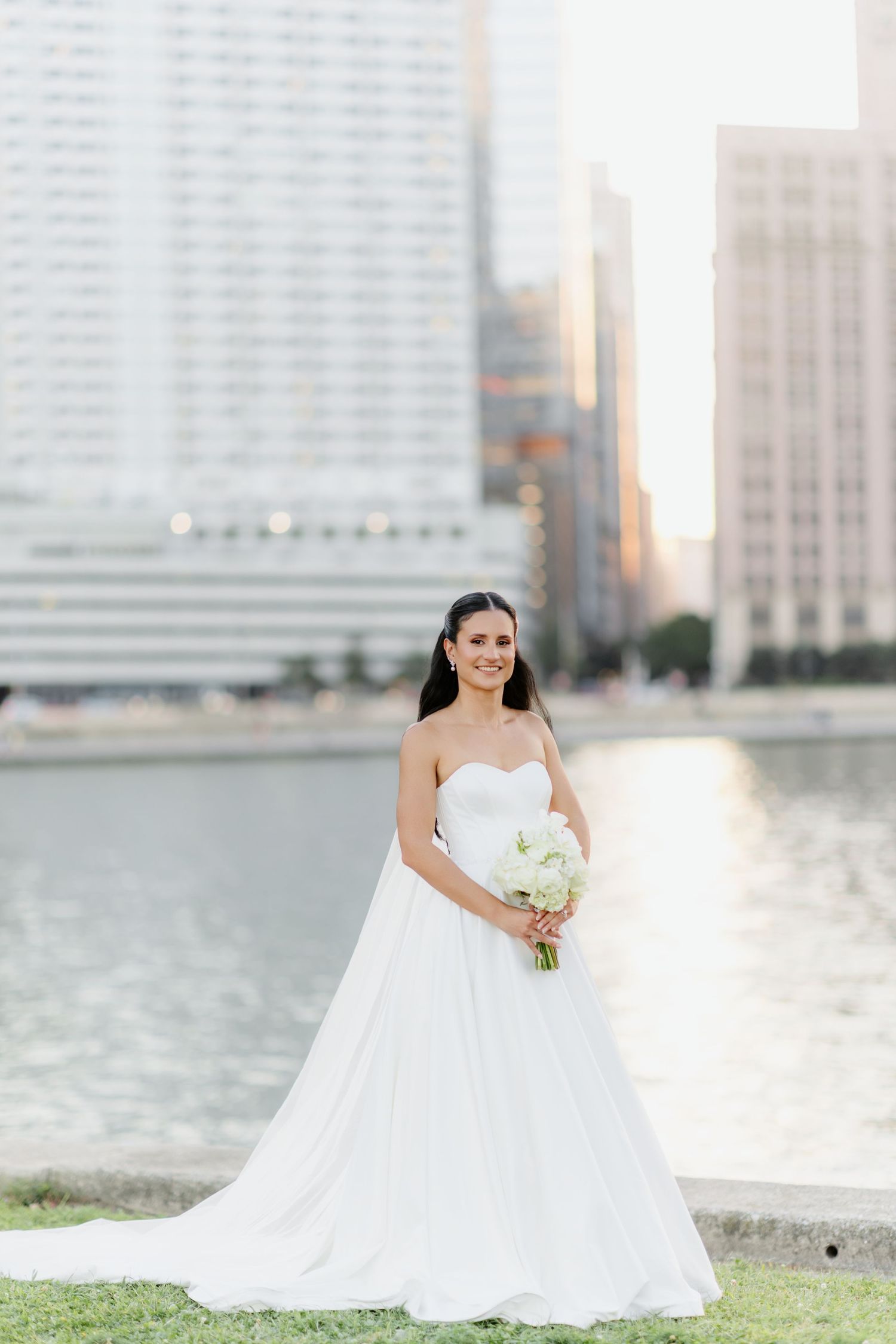 Bride in strapless white ballgown holding bouquet by urban waterfront at golden hour.