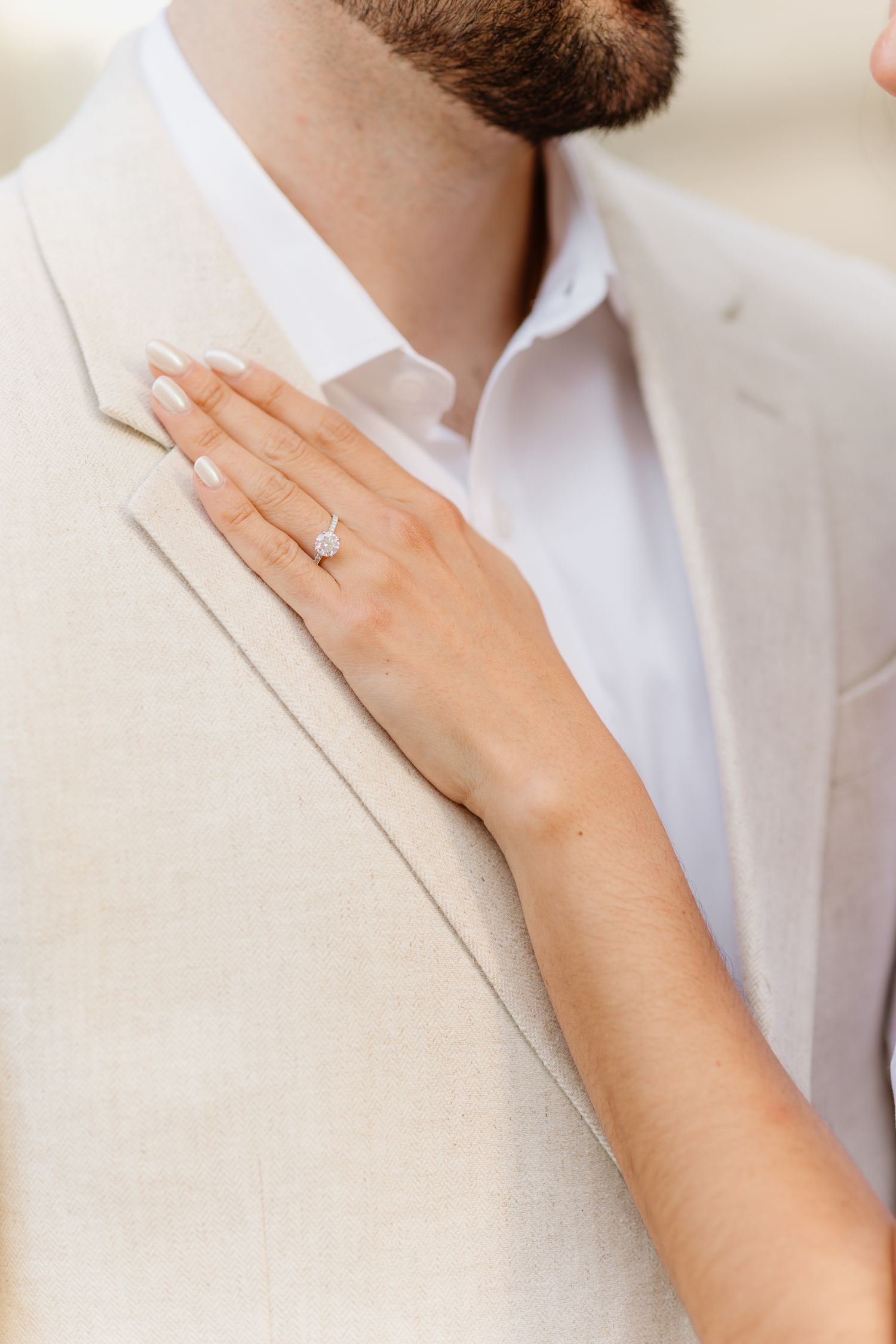 Bride's hand with diamond engagement ring resting on groom's beige linen suit jacket at wedding.