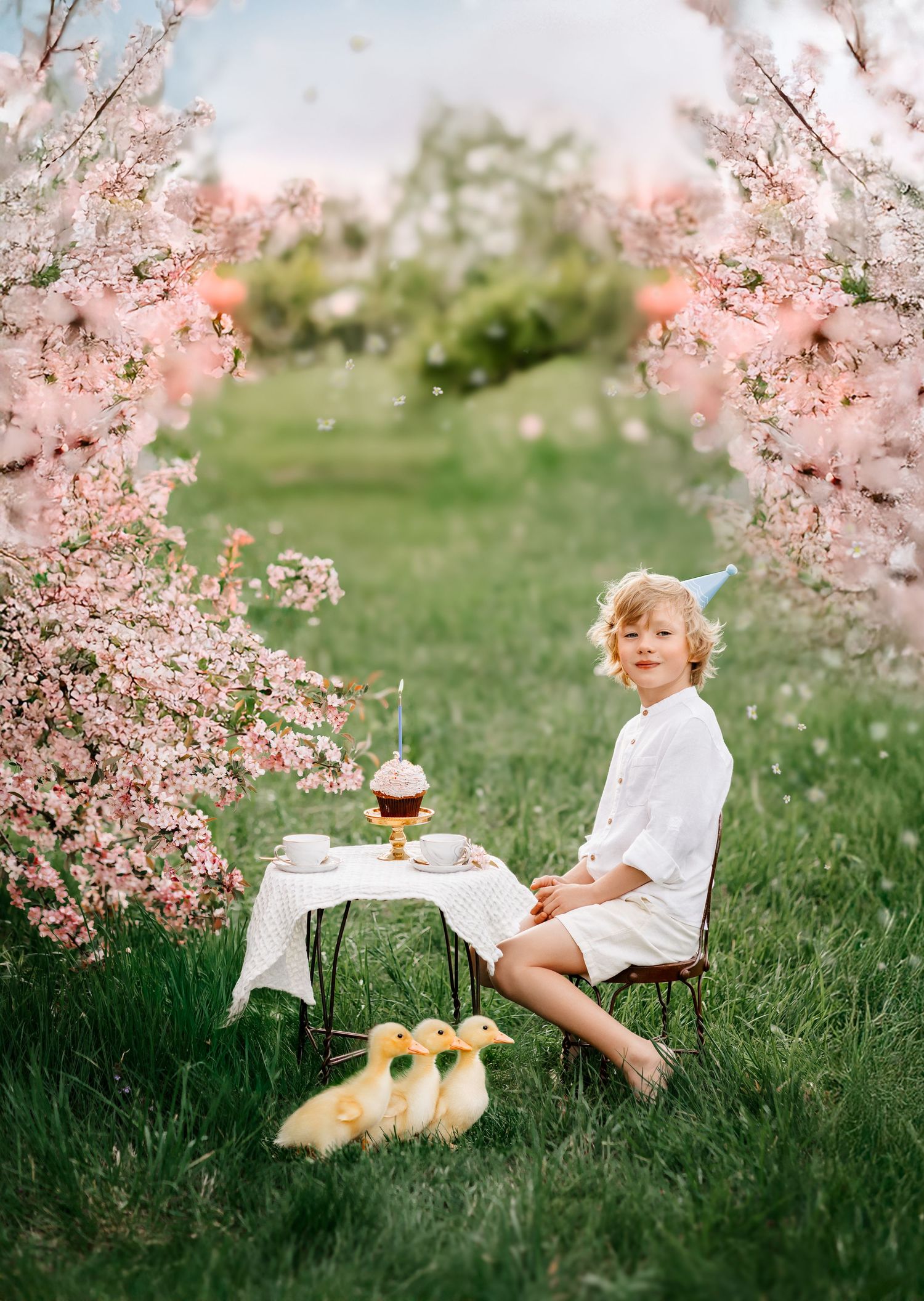 A child in white sits at a small table with ducklings beside blooming pink cherry trees during a birthday celebration outdoors.