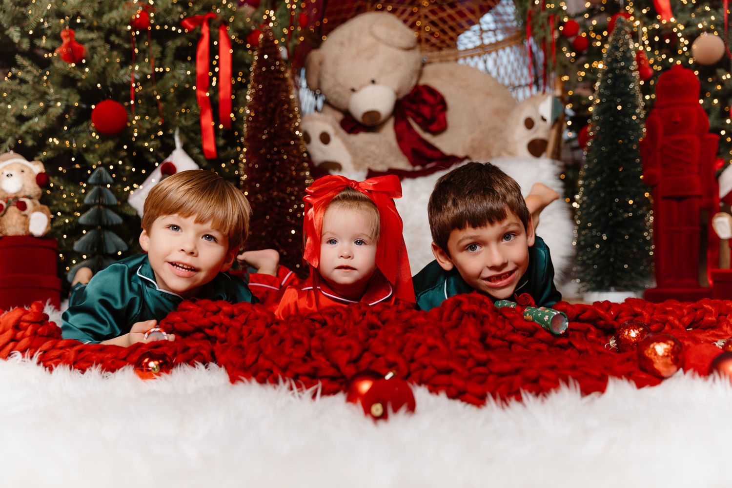 Three children pose on red and white blankets among Christmas trees, ornaments, and a teddy bear in a festive holiday scene.
