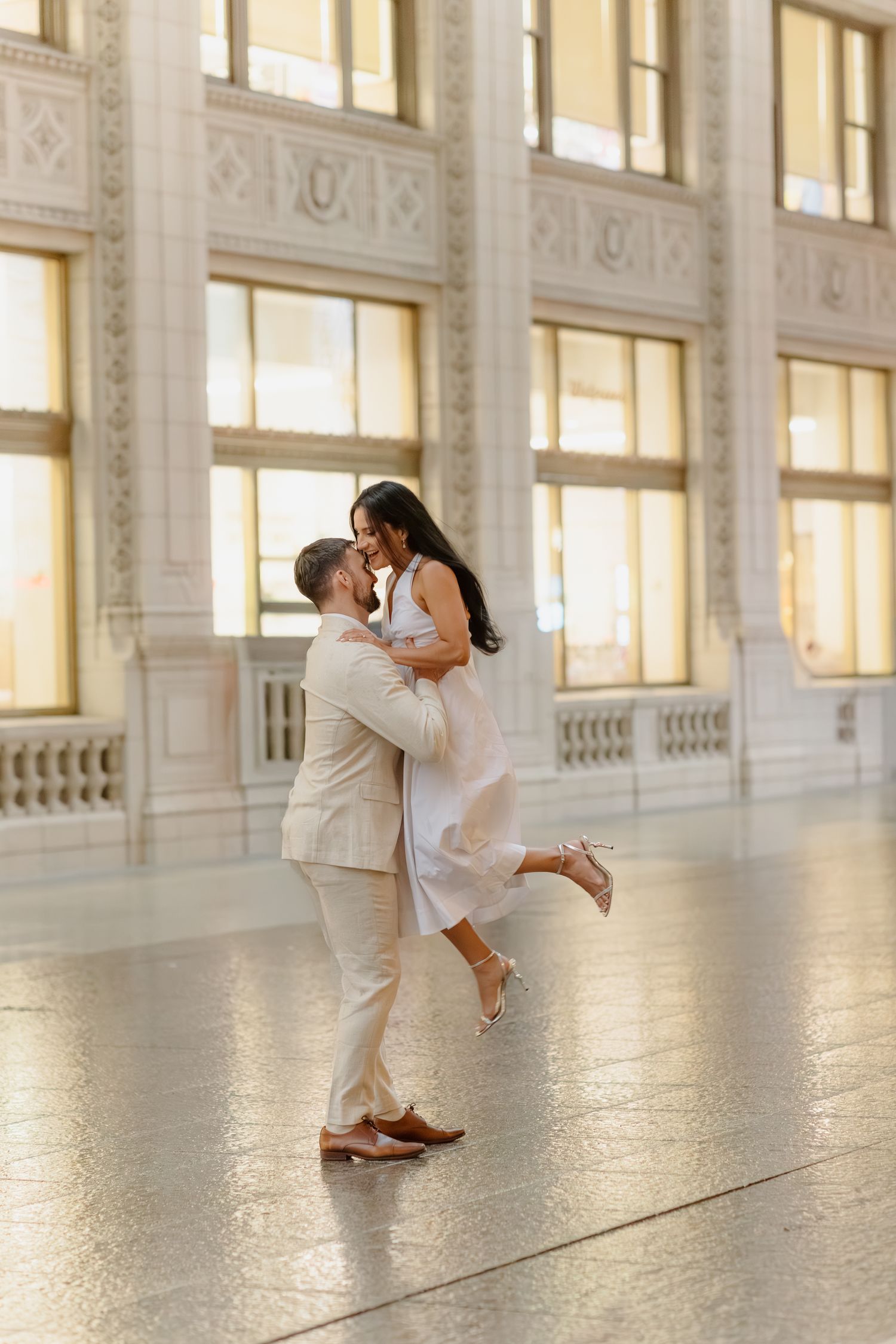 A couple shares a romantic embrace in an elegant grand hall, the man lifting his partner as she raises one heel.