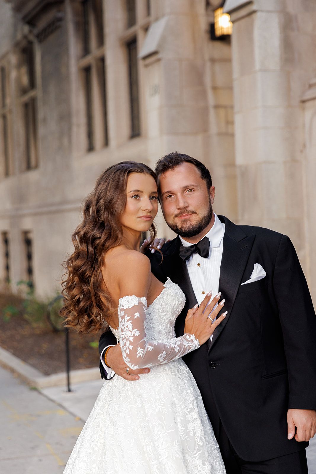 Bride in lace off-shoulder wedding gown embracing groom in black tuxedo outside a historic stone building.