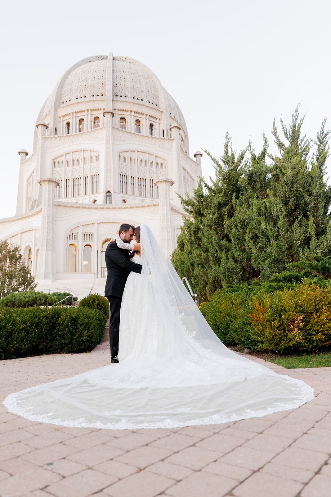 Wedding couple share romantic moment before ornate white Bahai Temple, bride's cathedral veil dramatically spread across ground.