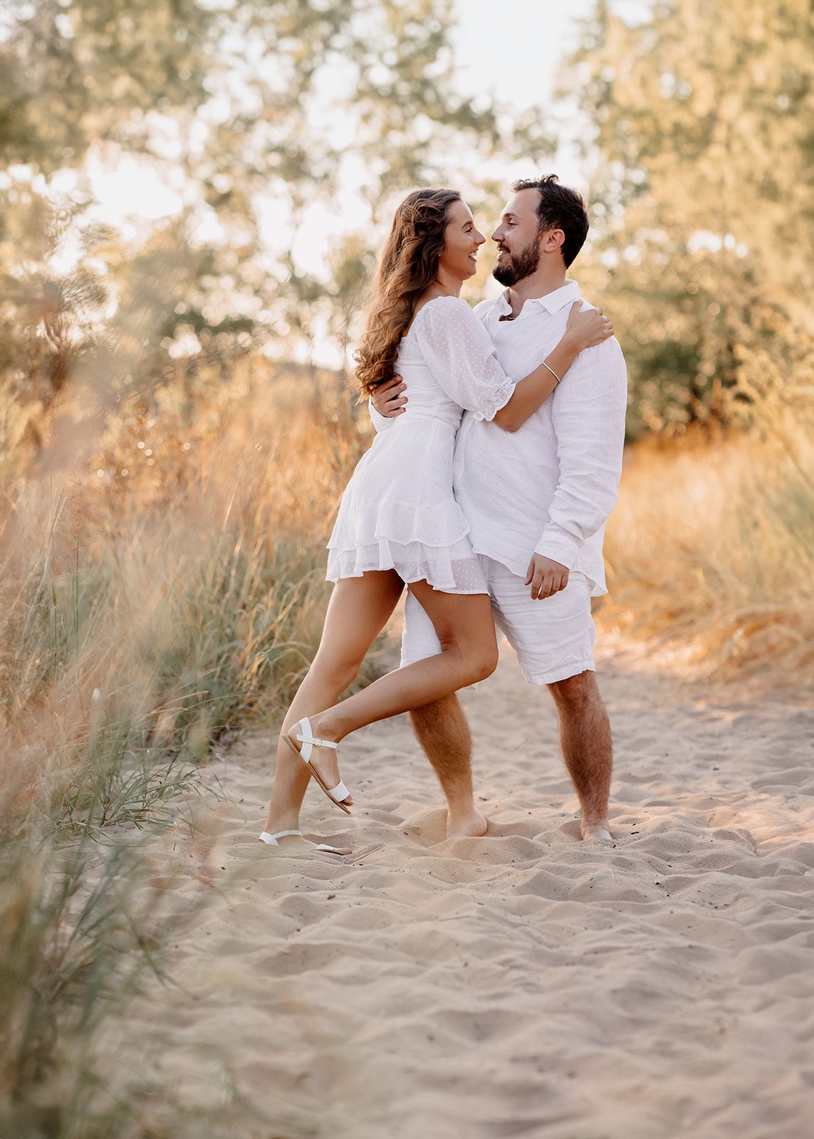 Couple in matching white outfits embracing on sandy beach path surrounded by golden grass and trees.