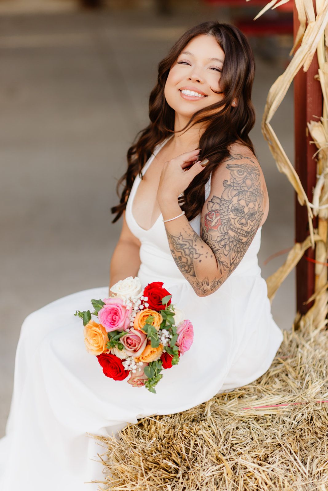 Tattooed bride in white gown sits on hay bale holding colorful red and pink rose wedding bouquet in barn.